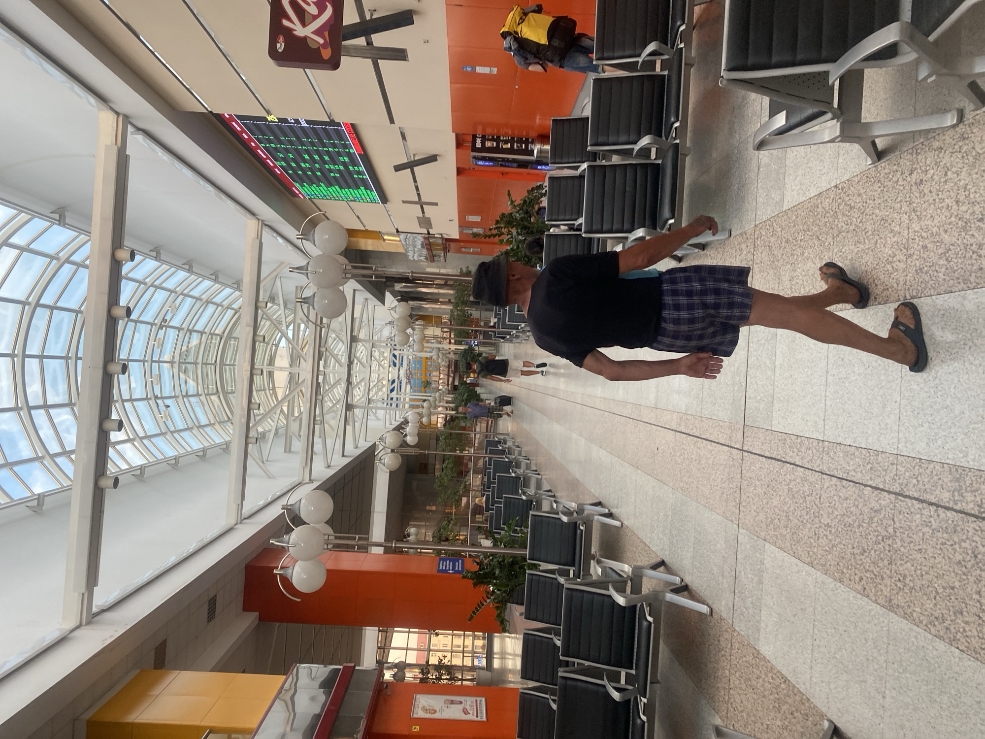 This is a wide-angle, eye-level shot of an indoor waiting area, likely an airport or bus station, with a glass ceiling. In the foreground, a man wearing a black t-shirt, plaid shorts, a black hat, and flip-flops walks away from the camera. He is in the center of a wide, tiled walkway. To his left and right are rows of empty black seats with metal frames.
Further down the walkway, a man in a blue shirt and dark shorts pulls a black suitcase on wheels. Another man, wearing a blue shirt and carrying a yellow backpack, walks away from the camera towards the right side of the image. In the background, a digital board displays green text and numbers. The walls are painted in orange and white. The overall impression is of a spacious, modern waiting area. The presence of a flight information board suggests this could be an airport in Minsk, Belarus, given the context.