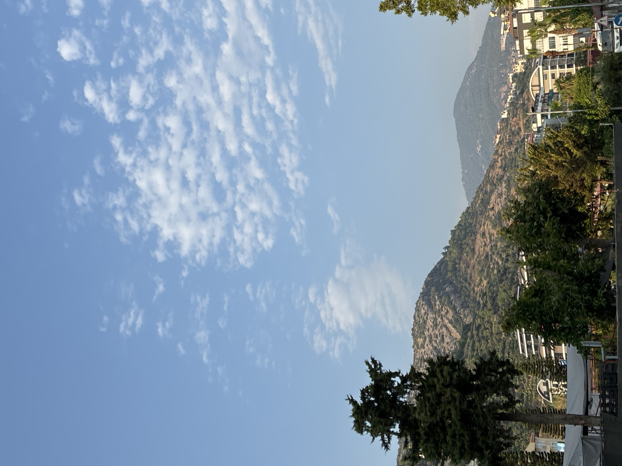 The image captures a view of Alanya, Turkey, dominated by a clear blue sky with scattered white clouds. In the foreground, a tall, dark green evergreen tree stands to the left. Behind it, the landscape transitions to hills covered in sparse vegetation and rocky outcrops. Further in the background, a town with several multi-story buildings, some with modern architecture featuring balconies and white facades, is nestled at the foot of the mountains. Green trees and smaller bushes are interspersed throughout the town and on the slopes of the hills. A road with some traffic signs is visible at the bottom right, and a white vehicle, possibly a truck, is partially seen. The overall scene depicts a sunny day in a coastal Mediterranean city, with a blend of natural mountainous terrain and urban development.