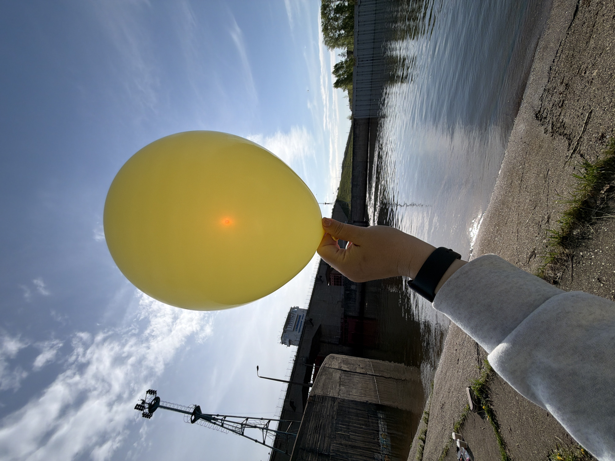 A person is holding a yellow balloon in front of the Chaikovsky gateway. The waterway is visible with sunlight reflecting on the surface. A concrete embankment with vegetation lines the side. The sky is blue with scattered clouds. A section of the lock structure and a crane are also visible. The person's arm, wearing a grey sweatshirt and black wristwatch, is in the lower right corner.