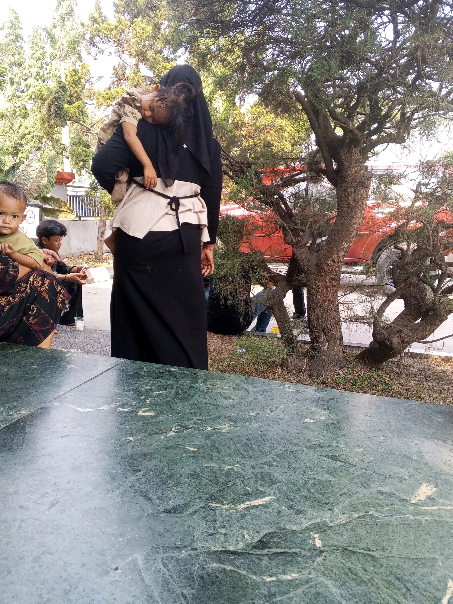 In Ciruas, Indonesia, an outdoor scene depicts a woman in a black hijab and long black skirt carrying a sleeping child. The child is nestled against the woman's shoulder, wearing a tan and beige outfit. To the left of the woman, a young child with light skin and dark hair sits, looking towards the camera. Another child, appearing to be male, is seated further back, holding a small piece of food. In the background, a red vehicle is partially visible behind a tree with dense, dark branches. The ground is paved, and there is green grass and vegetation. A green marble table occupies the foreground, obscuring the lower portion of the scene.