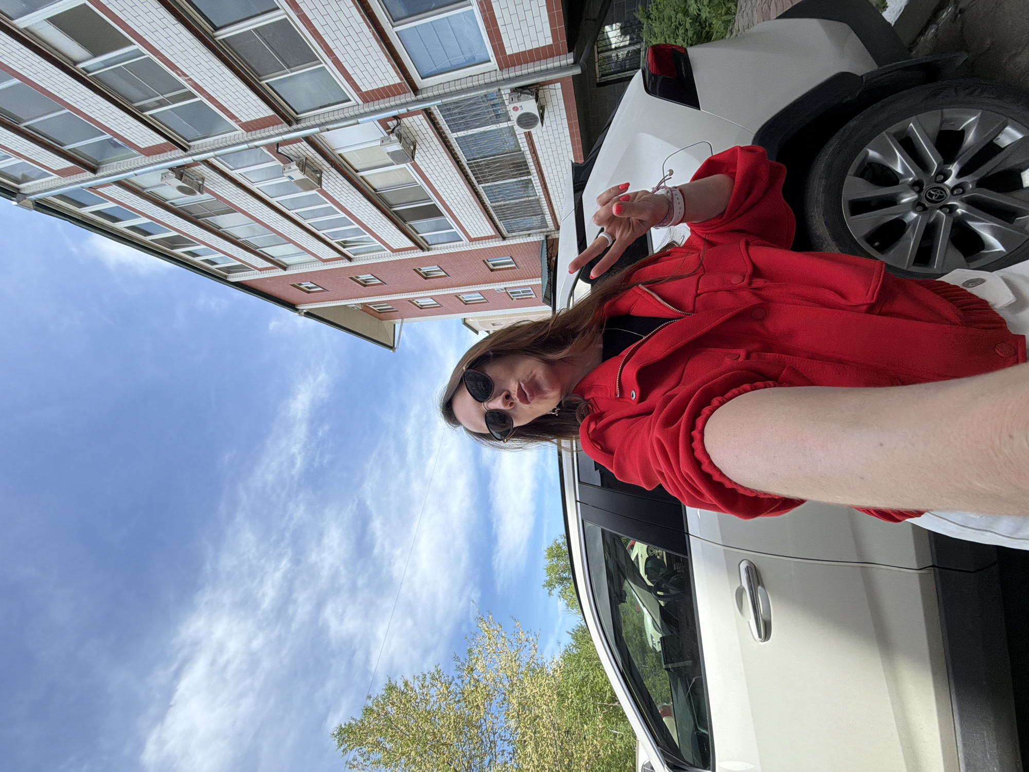 A woman wearing a red jacket and sunglasses poses for a selfie next to a white car. She is making a peace sign with her right hand. In the background is a brick building with white trim and numerous windows. The sky is blue with wispy clouds. There are trees in the lower left corner of the image. The location is Birobidzhan, Russia.