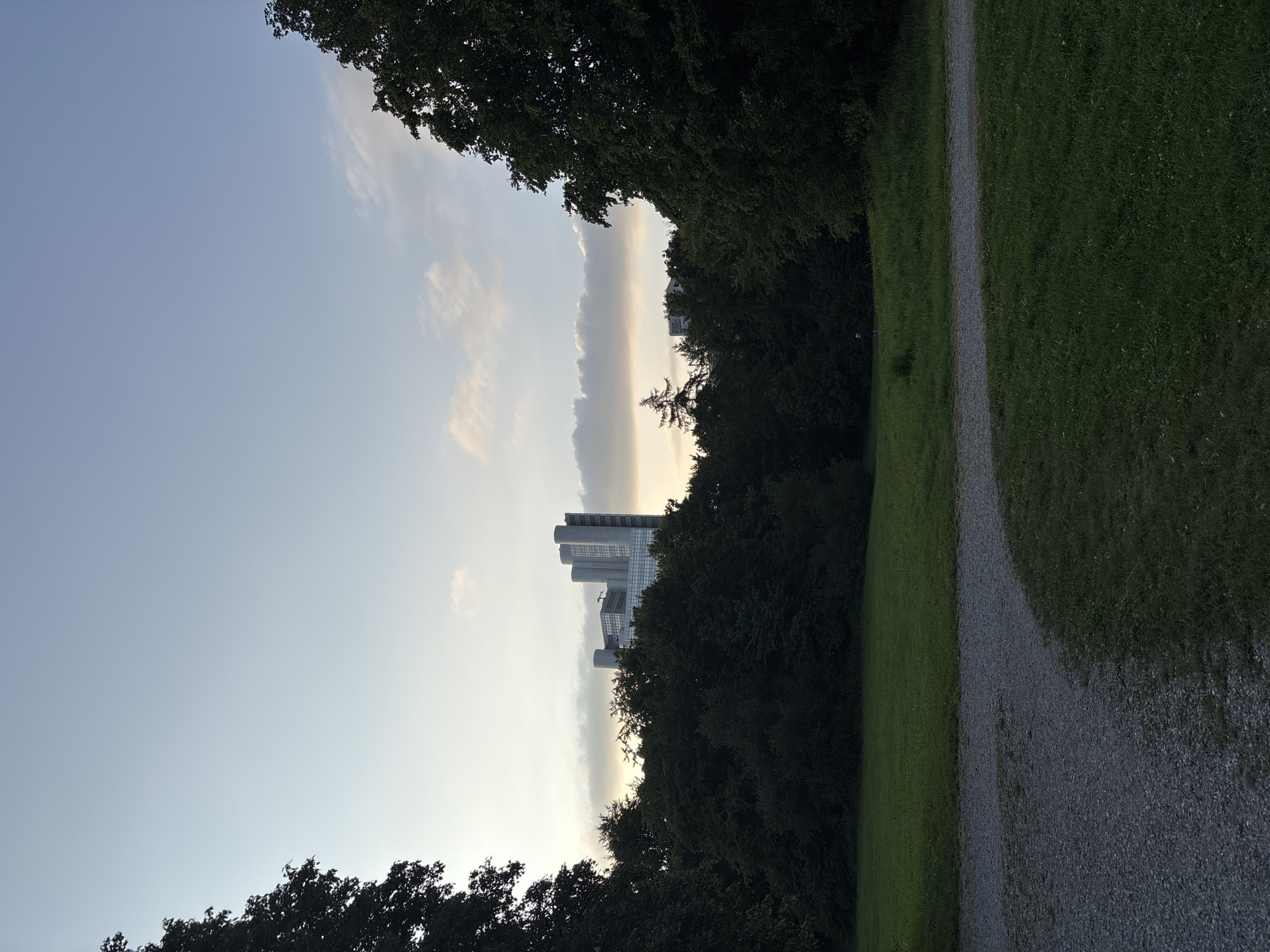 This image captures a serene park scene in Munich, Germany, likely during the late afternoon or early evening. In the background, a modern, multi-story building with a distinctive architectural style stands tall, peeking through a dense canopy of dark green trees. The building appears to be a corporate office or residential complex, characterized by its glass facade and multiple cylindrical towers.
In the foreground, a gravel path curves gently through a lush green lawn, suggesting a well-maintained public space. The path is a mix of light gray and dark gray stones. The lawn is neatly trimmed, with scattered white wildflowers adding a touch of natural beauty. The sky above is a soft, pale blue, transitioning into a warmer hue near the horizon, indicating the presence of clouds that are illuminated by the setting sun.
On the far right of the image, a large, mature tree with dark green foliage partially frames the scene. On the far left, another large tree is visible, its silhouette darker against the sky. In the middle ground, a solitary figure, a person with dark clothing, is walking on the lawn, adding a sense of scale and human presence to the otherwise tranquil landscape. The overall atmosphere is peaceful and idyllic, typical of a summer evening in a city park.