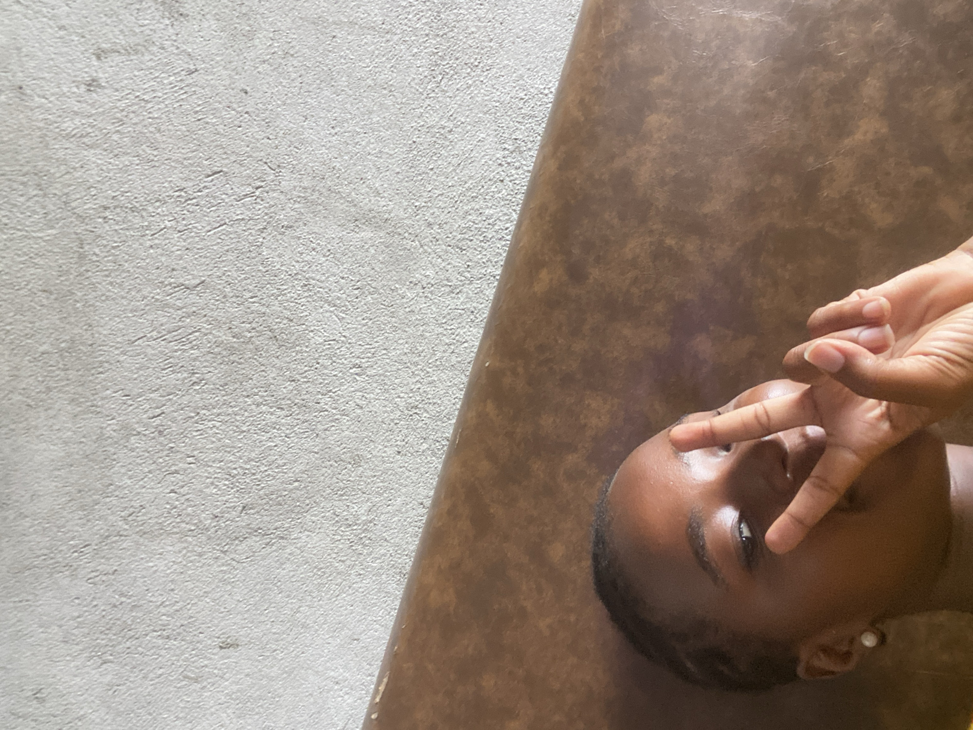 An upward-angled, medium shot shows a dark-skinned person with short hair, laying on a brown surface. The person's right hand is raised, fingers splayed over their face. The left side of the frame features a textured, light gray surface. No city or landmarks visible. No evidence to suggest the city of Dar es-Salaam, Tanzania.
