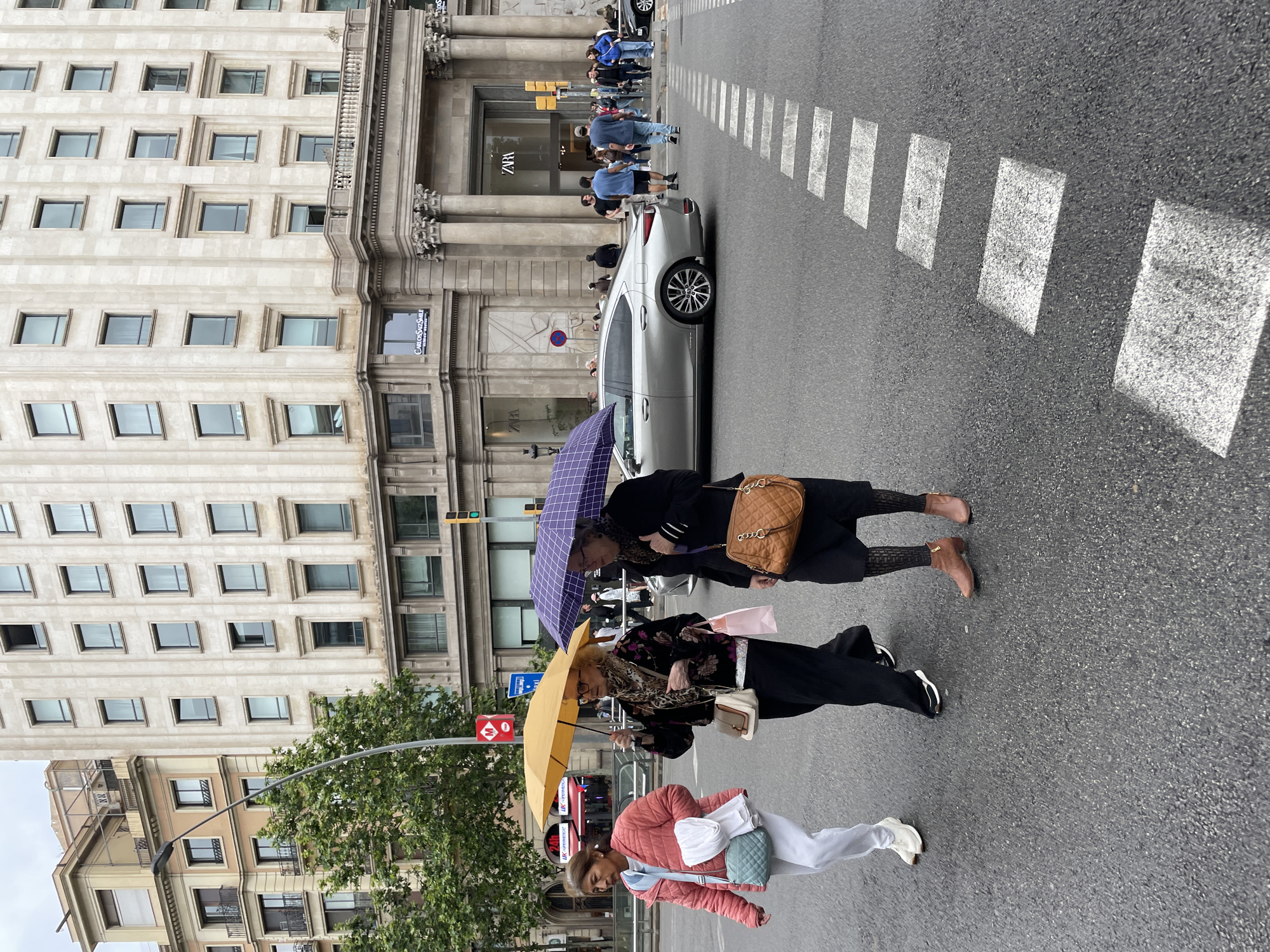 Three women are walking across a street in Barcelona, Spain. The women are walking on a crosswalk, and a silver car is stopped behind them. A Zara store is visible in the background behind the car, and other people are standing in front of the store. A building is visible on the left side of the image. The women are holding umbrellas. The first woman is wearing a pink jacket and white pants. The second woman is wearing a floral top and black pants, and is holding a yellow umbrella. The third woman is wearing a black jacket and black pants, and is holding a purple umbrella and a brown purse.