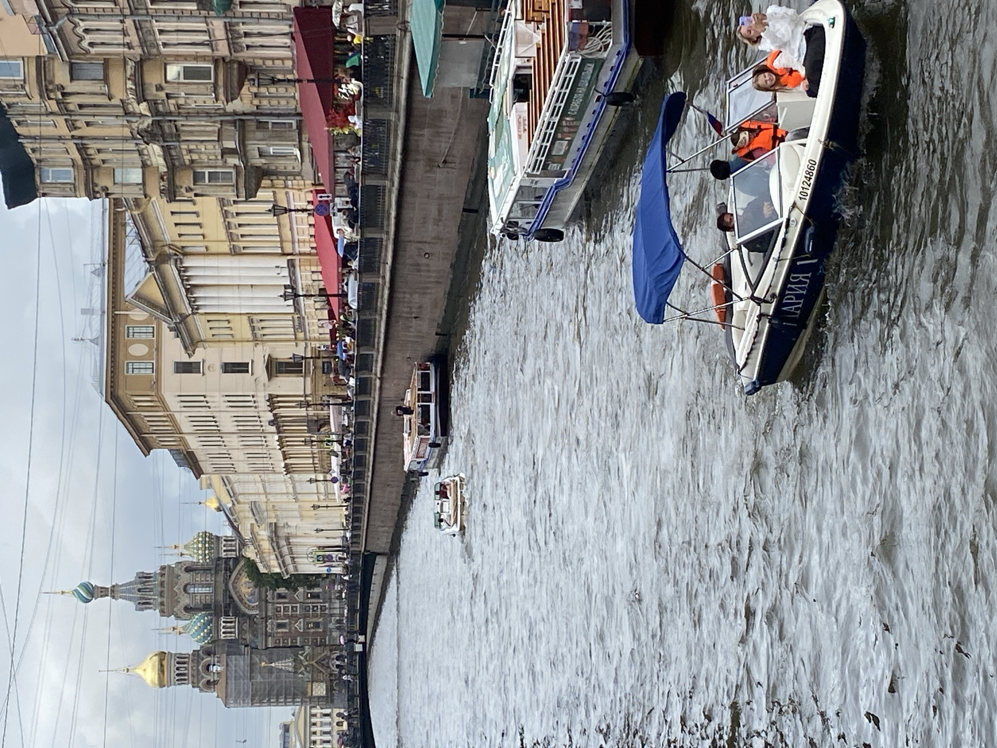 The image depicts a cloudy day in Saint Petersburg, Russia, with the Church of the Savior on Spilled Blood prominently visible in the background. Several boats are navigating a canal that runs alongside a row of buildings with awnings. In the foreground, a small motorboat with a blue canopy carries five people, four of whom are wearing orange life vests. One person in the boat is taking a photograph with a smartphone. Another larger boat, likely a tour boat, is docked on the right side of the canal, and a smaller white boat is moving away from the foreground. The canal is bordered by a stone embankment with a railing, behind which people are walking and sitting at outdoor cafes. The architecture of the buildings is characteristic of Saint Petersburg, with a mix of classical and more ornate styles. The water in the canal appears choppy.