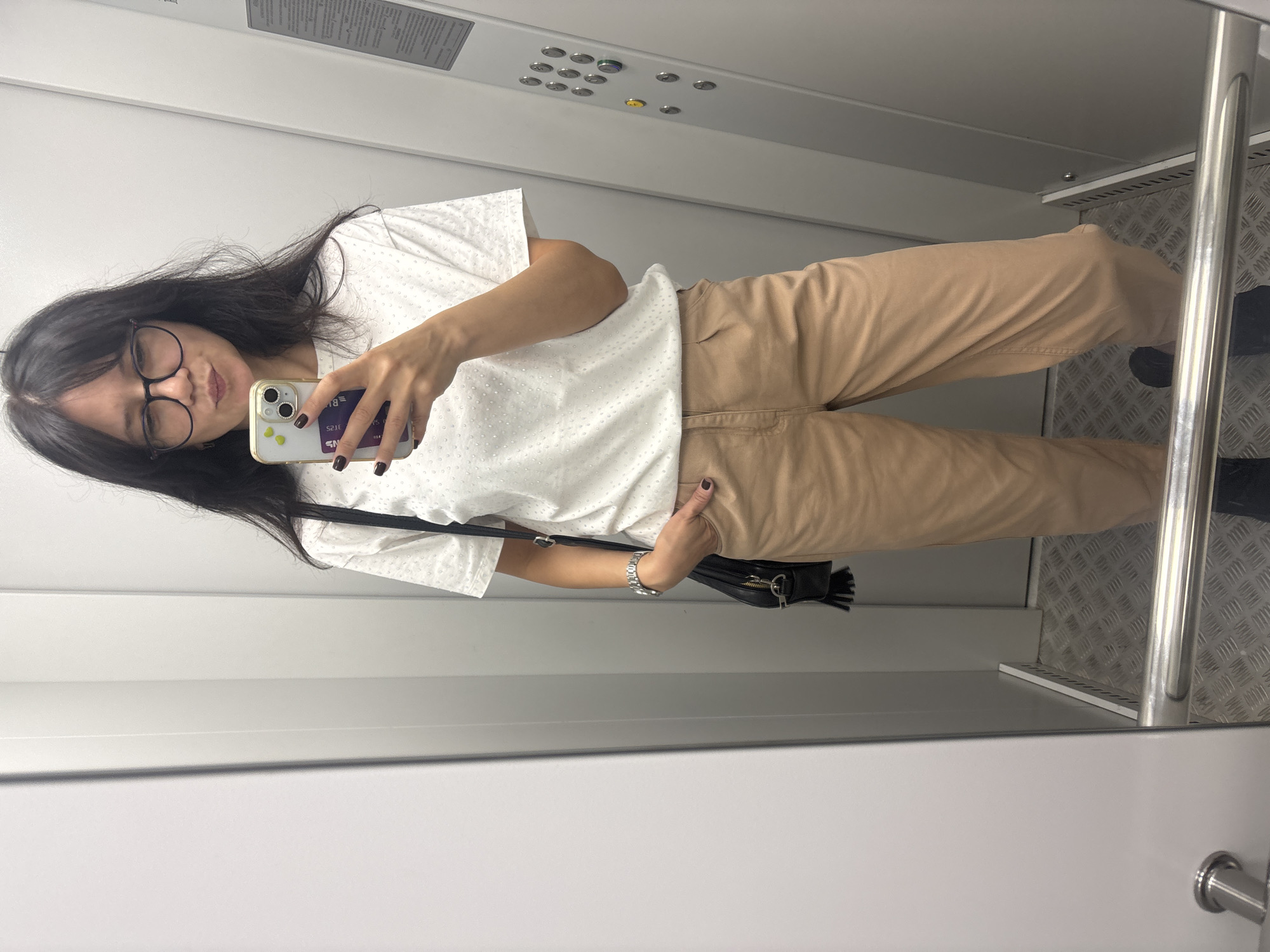 A young woman with dark hair and glasses is taking a mirror selfie in an elevator. She is wearing a white t-shirt with a subtle pattern, tan pants, and a black purse with tassels. Her right hand is in her pocket, and she is holding her phone in her left hand. The elevator appears to be modern, with metal walls and floor, and an elevator control panel on the right side. The image does not provide enough information to determine its location in the city of Chaikovsky, Russia.