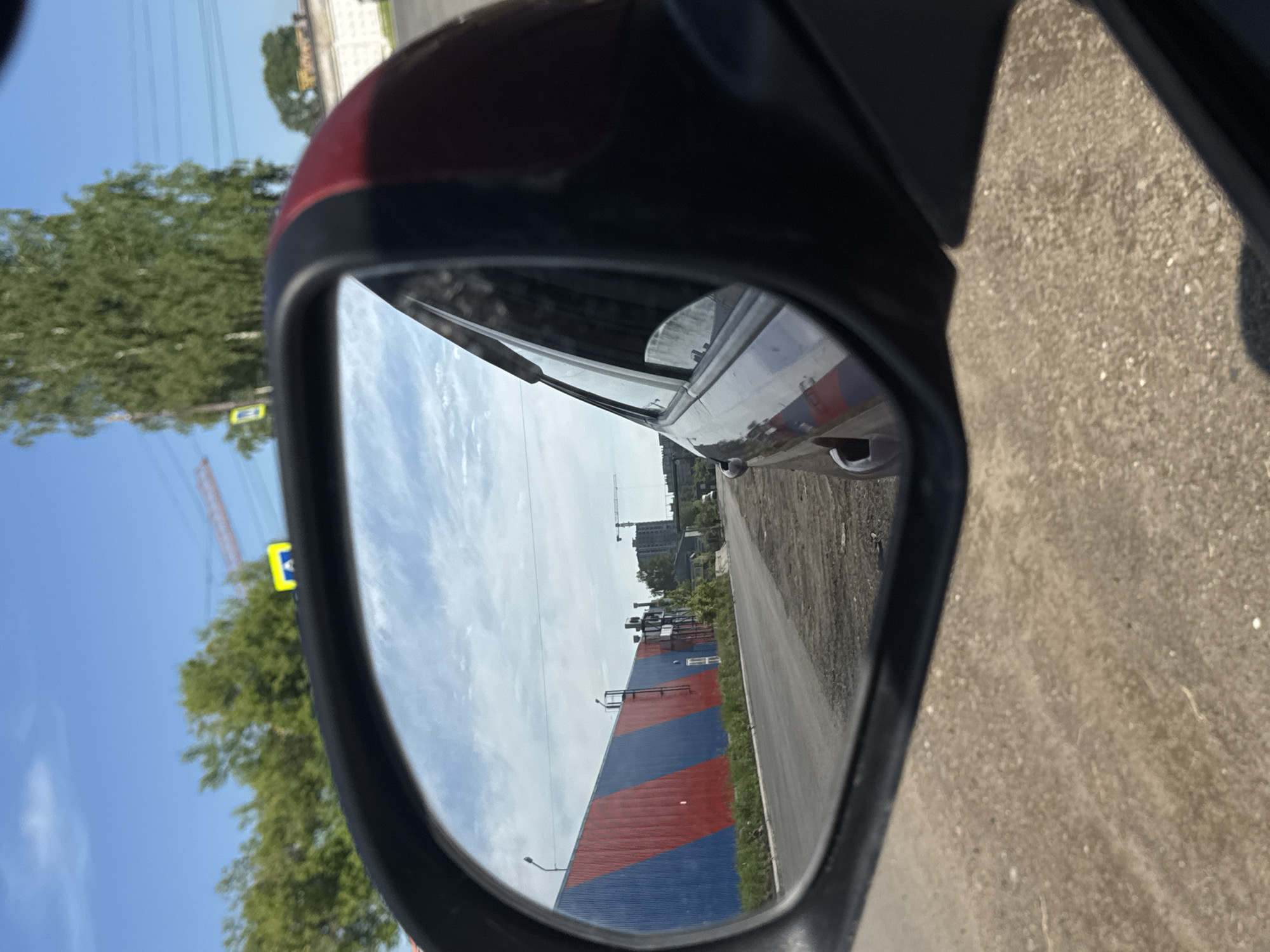 The image presents a view from inside a vehicle, looking into its red and black side mirror, on a bright, sunny day in Chaikovsky, Russia. The top edge of the mirror's casing is red, matching the car's body, while the rest of the mirror's frame and the immediate car interior visible are black. Textured tan-colored pavement or ground is visible outside the car, to the far right.
The reflection in the side mirror captures a clear blue sky with some wispy white clouds across the top. Below the horizon line, a paved road or pathway stretches into the distance, running parallel to a large, prominent building. This building features a distinctive pattern of wide, diagonal stripes in alternating red and blue colors. Beyond this striped building, in the background, several multi-story structures and what appears to be a construction crane are visible, indicating an urban or industrial setting.
Outside the mirror's reflection, to the left of the mirror, vibrant green trees are abundant against the bright blue sky. Overhead, multiple power lines crisscross the sky. Partially visible amidst the trees and power lines is a yellow sign bearing a dark, capital letter, resembling an 'E'. The scene depicts a daytime environment, possibly in an industrial or developing area, characterized by clear weather conditions.