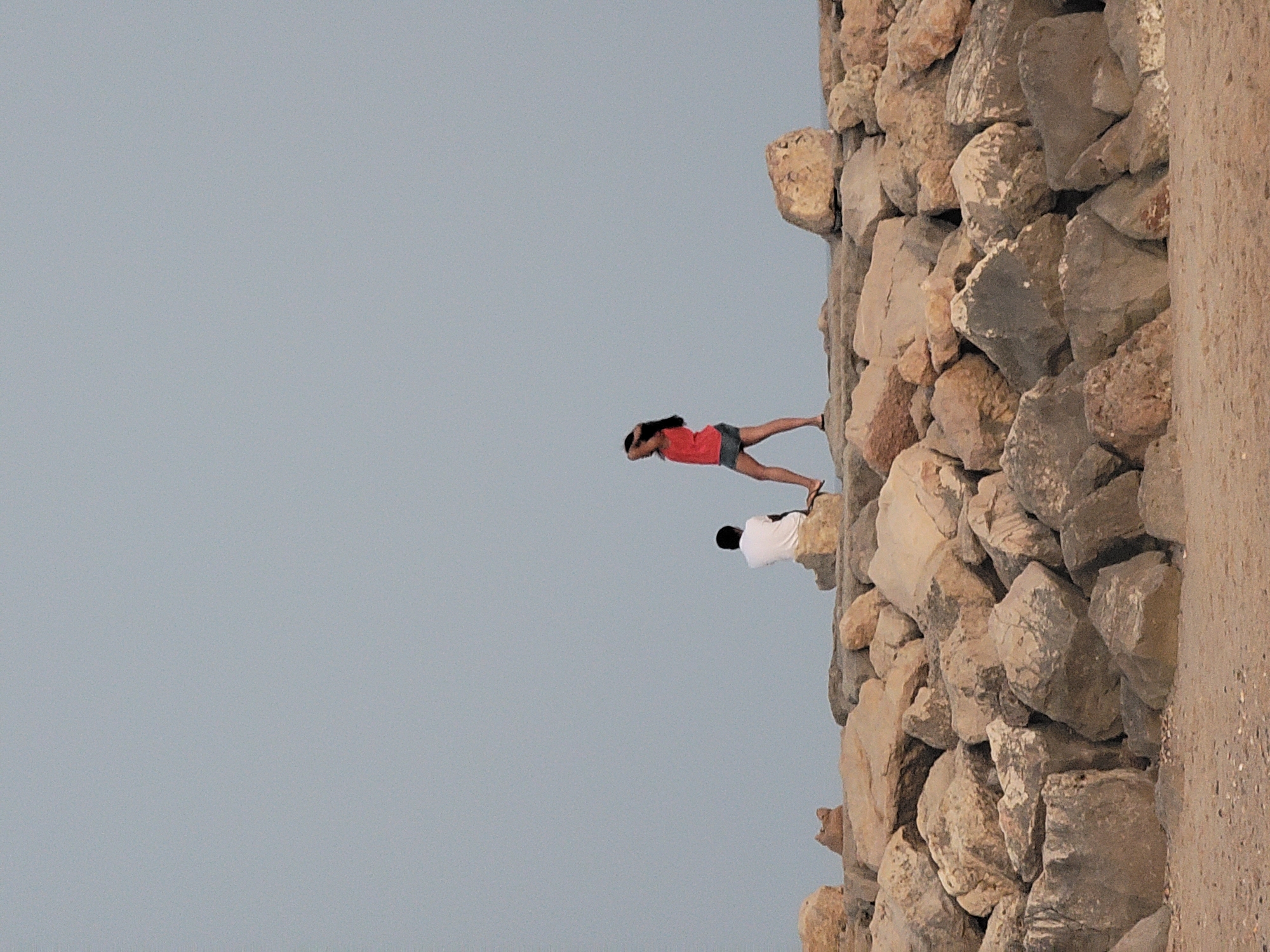 Two individuals are positioned on a rocky embankment, likely part of the Quarteira coastline in Portugal. The person on the left, viewed from the back, is seated on a rock wearing a white shirt. To their right and slightly further away, another person stands, facing left. This individual is wearing a reddish-orange sleeveless top and denim shorts, with their right hand touching their hair. The background consists of a clear, pale blue sky. The foreground is composed of a sandy beach. The embankment itself is constructed from numerous large, irregular rocks of varying sizes and colors, predominantly in shades of grey and brown.