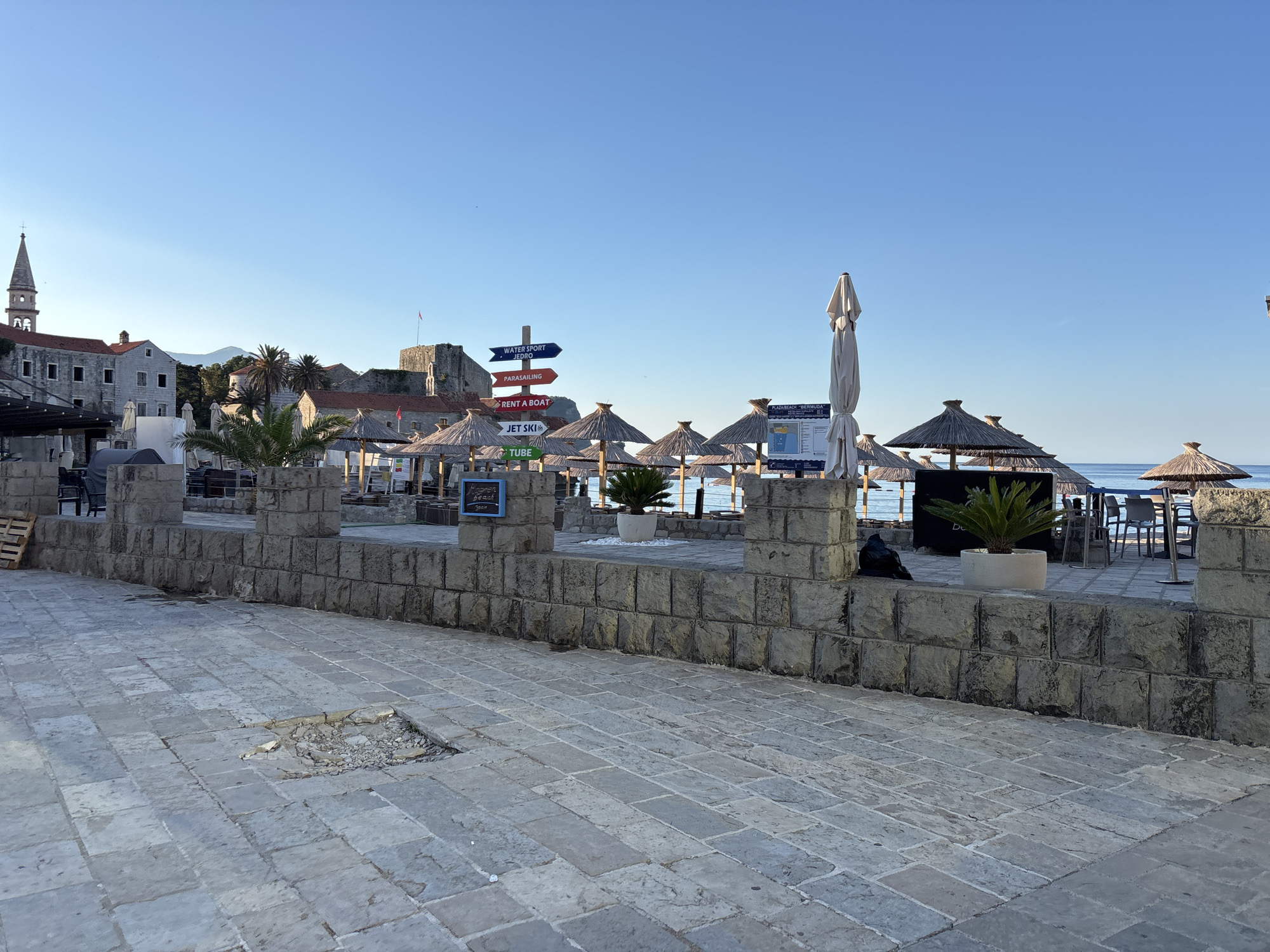 A coastal scene is captured under a clear blue sky. In the foreground, a paved walkway constructed of large, rectangular stone slabs is visible, with a section of broken paving stones revealing gravel underneath. A low, thick stone wall with a rough, blocky texture borders the walkway. Behind the wall, a sandy area with numerous straw-thatched umbrellas is set up, with some closed and others open. Several potted palm plants are scattered amongst the umbrellas. In the distance, the sea is calm and blue.
To the left, a collection of stone buildings with red tile roofs is present, including a tall bell tower with a cross on top. A medieval-style fortress or castle with stone walls and a red flag flying from its ramparts is also visible. A directional signpost with multiple arrows indicating "WATER SPORT," "JET SKI," "RENT A BOAT," and "PARASAILING" stands near the center of the image. In the background, a few palm trees punctuate the landscape, and distant mountains are faintly visible on the horizon. The scene appears to be a tourist area, possibly a promenade or beach entrance, in a historic coastal town. There are no people visible in the frame.