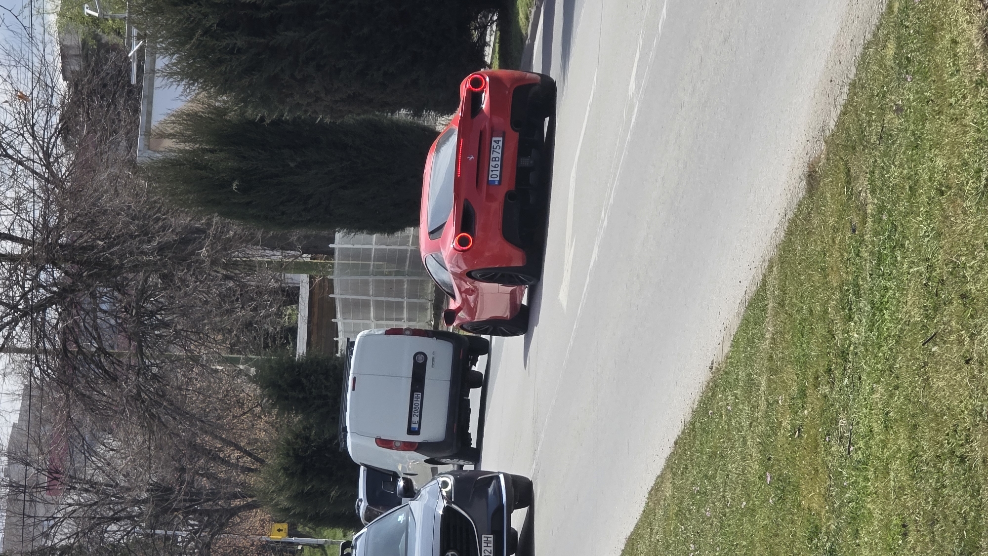 The image shows a street scene with several vehicles in Blagoevgrad, Bulgaria. At the front of the line is a dark-colored Mazda SUV with the license plate "EB32HH." Behind the Mazda is a white Fiat Doblo van with the license plate "E2080HH." A red Ferrari is directly ahead of the van, displaying the license plate "016B754." The vehicles are parked on a gray asphalt road next to a grassy area. Trees are visible on the left side of the image. A building is visible between the trees and shrubbery.