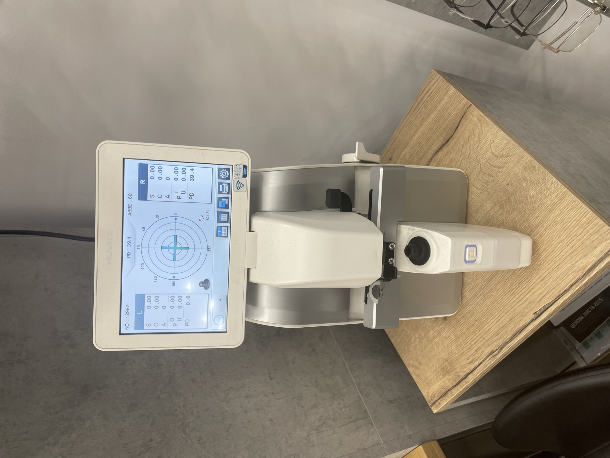 The image presents an autorefractor, a diagnostic instrument used in ophthalmology to measure the refractive error of the eye. The device, likely a model from Huvitz, is positioned on a light-colored wooden surface resembling a countertop or table. The screen displays readings for both left (L) and right (R) eyes, with zero values for Sphere (S), Cylinder (C), Axis (A), Prism (P), and Prism Up (U). The Pupillary Distance (PD) is measured at 39.4 for the right eye and 0.0 for the left eye. The ABBE number is set to 60.
The environment suggests a clinical setting, perhaps an optometry office or eye clinic. The presence of eyeglass frames and a chair further supports this context.
"ЦЕНОВА ЛИСТА 2025" appears to be Bulgarian text, indicating that the photo might have been taken in Bulgaria. Without further information, it's impossible to determine the exact location within Sofia.