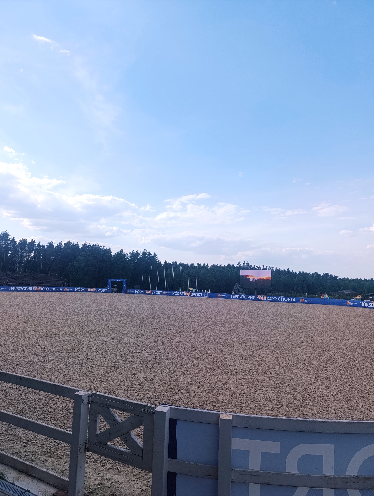 The image captures an outdoor horse-riding arena, filled with sand. Along the perimeter of the arena, banners with Russian text and "HORSEKA SPORT" are visible. In the background, a dark green line of trees separates the arena from the sky. A large screen is visible in the distance, possibly displaying event information. A wooden and blue fence is located on the front left of the image. The sky is a light blue with scattered clouds. The location is the city of Ivanovskoe, Russia, as indicated by information available online.
