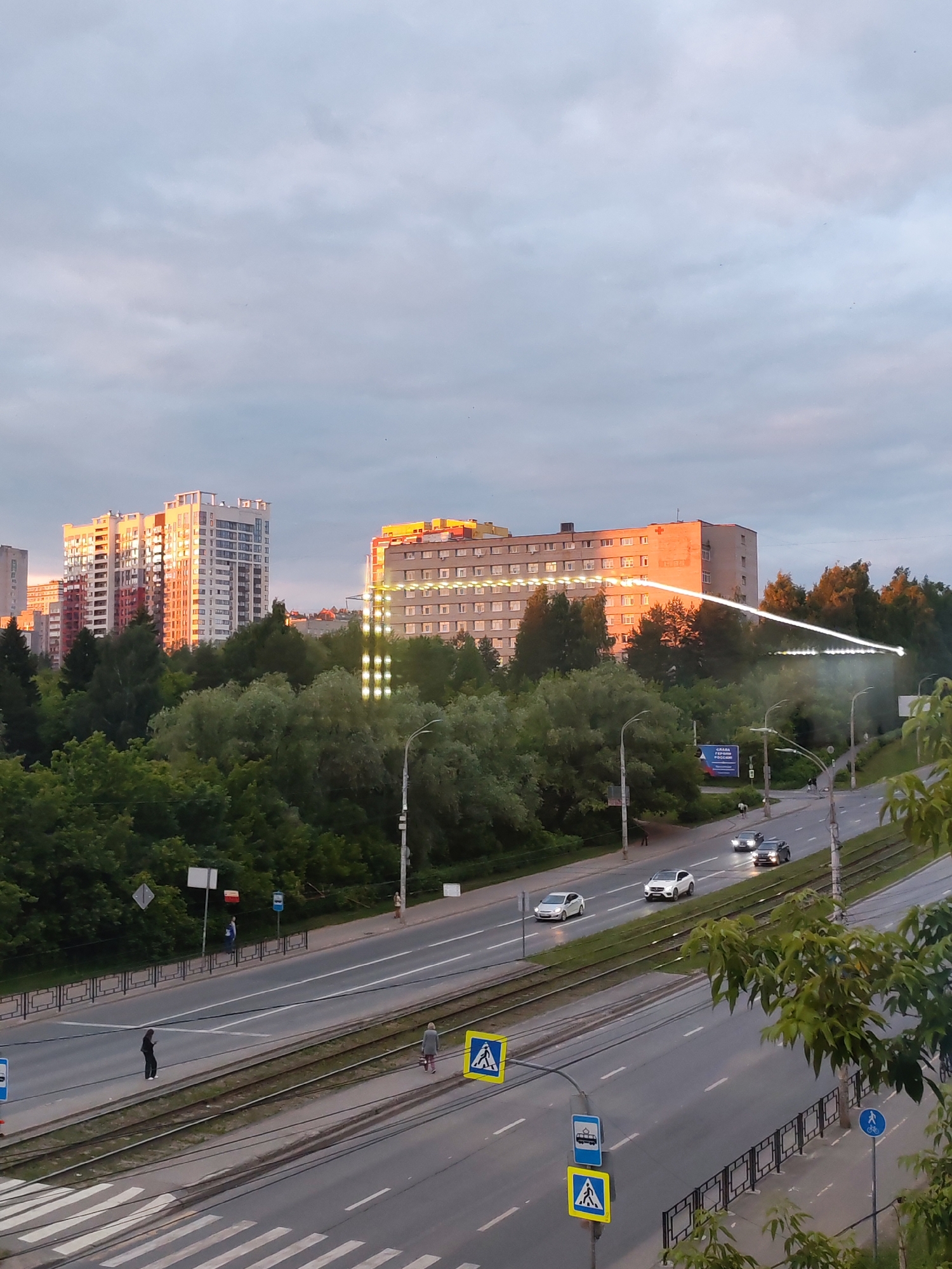 The image depicts a panoramic view of a city street in Izhevsk, Russia, during what appears to be late afternoon or early evening. The sky is overcast with diffused sunlight casting a warm glow on some of the buildings. In the background, several modern apartment buildings are visible, some with strikingly illuminated facades.
A prominent feature in the mid-ground is a long, multi-story building with a distinctive pattern of lights, possibly a medical facility or a research institute, illuminated by the setting sun. This building is flanked by dense green trees, indicating a park or natural reserve within the urban landscape.
The foreground shows a multi-lane road with light traffic. Cars are driving along the road, and tram tracks are present alongside the lanes. Pedestrians are visible on the sidewalk, some waiting at bus stops, and others crossing the street at a marked crosswalk. Traffic signs and streetlights are also present, contributing to the urban setting. The overall impression is of a typical city scene in Izhevsk, Russia, with a blend of modern architecture, green spaces, and daily urban activities.