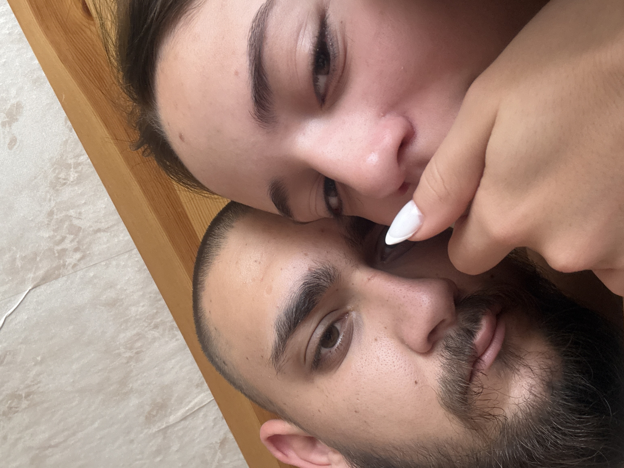 A close-up photograph captures two individuals, a woman and a man, in an intimate pose. The woman is positioned above the man, her face partially obscuring his. Her hand, with a white fingernail, is visible near his face. The background includes what appears to be wood grain and a textured surface, possibly a wall or bedding. The photo's orientation suggests it was taken from an unconventional angle. No evidence or indication of the location of Staro Selo, Bulgaria, is found within the image.