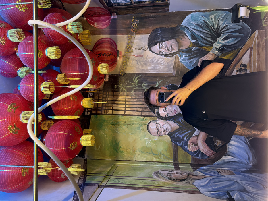 This image captures an interior scene, likely within a restroom, at a location in Canggu, Indonesia. The upper half of the frame is dominated by numerous red spherical Chinese lanterns adorned with golden tassels, suspended from the ceiling. White, flexible tube-like light fixtures or decorative elements are horizontally strung across the space, interweaving with the lanterns.
The background wall features an elaborate mural depicting four notable figures in an East Asian-style setting with traditional architecture and bamboo foliage. From left to right, the painted individuals resemble Kurt Cobain, a smiling Chester Bennington wearing glasses and holding a cup, Amy Winehouse looking towards Bennington, and John Lennon wearing glasses and a blue jacket. Mirrored, vertical text "tea & love" is visible on the mural behind the figures on the right.
In the foreground, a male subject is taking a mirror selfie. He is seen from the chest up, wearing a black short-sleeved t-shirt with a small red and white logo on the left chest. His face is obscured by a black smartphone, held by his right hand, which displays a triple-lens camera module. A tattoo is visible on his right forearm. On the far right, below the mural, a white wall-mounted toilet paper dispenser with a roll is present.