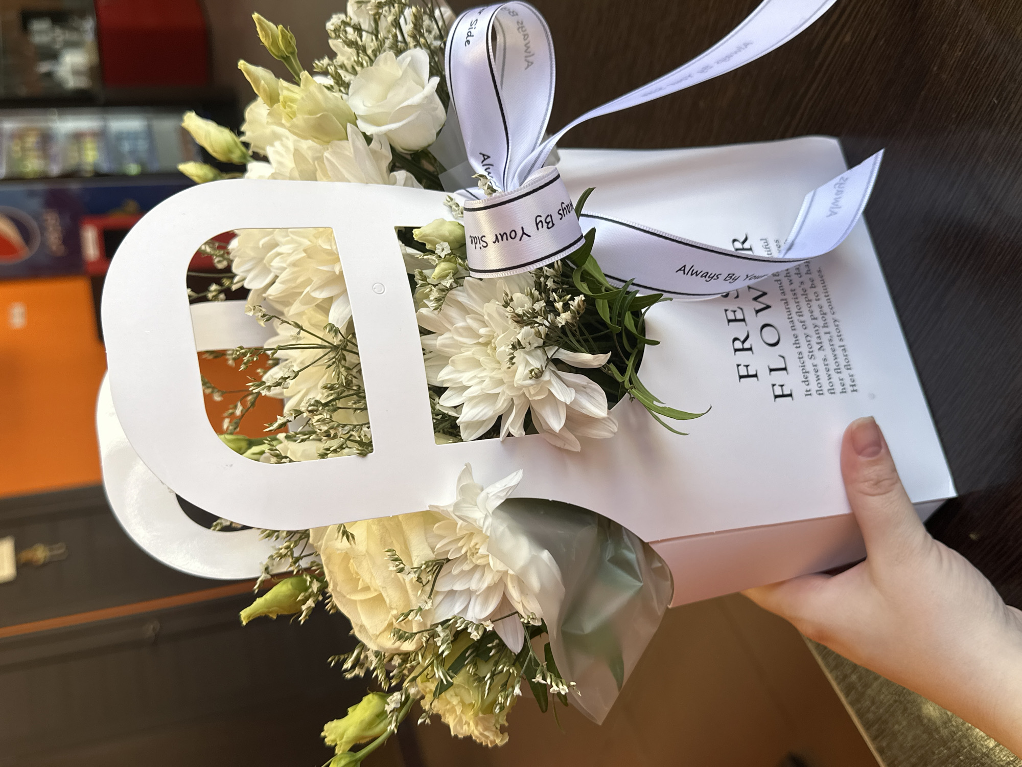 The image shows a white floral arrangement in a box. The box has cut-out letters in the shape of "FRESH FLOWERS" on the front. A white ribbon with black writing that reads "Always By Your Side" is tied around the bouquet. The arrangement consists of white roses and chrysanthemums with some green foliage. A hand is holding the box, and the background appears to be a table surface indoors.