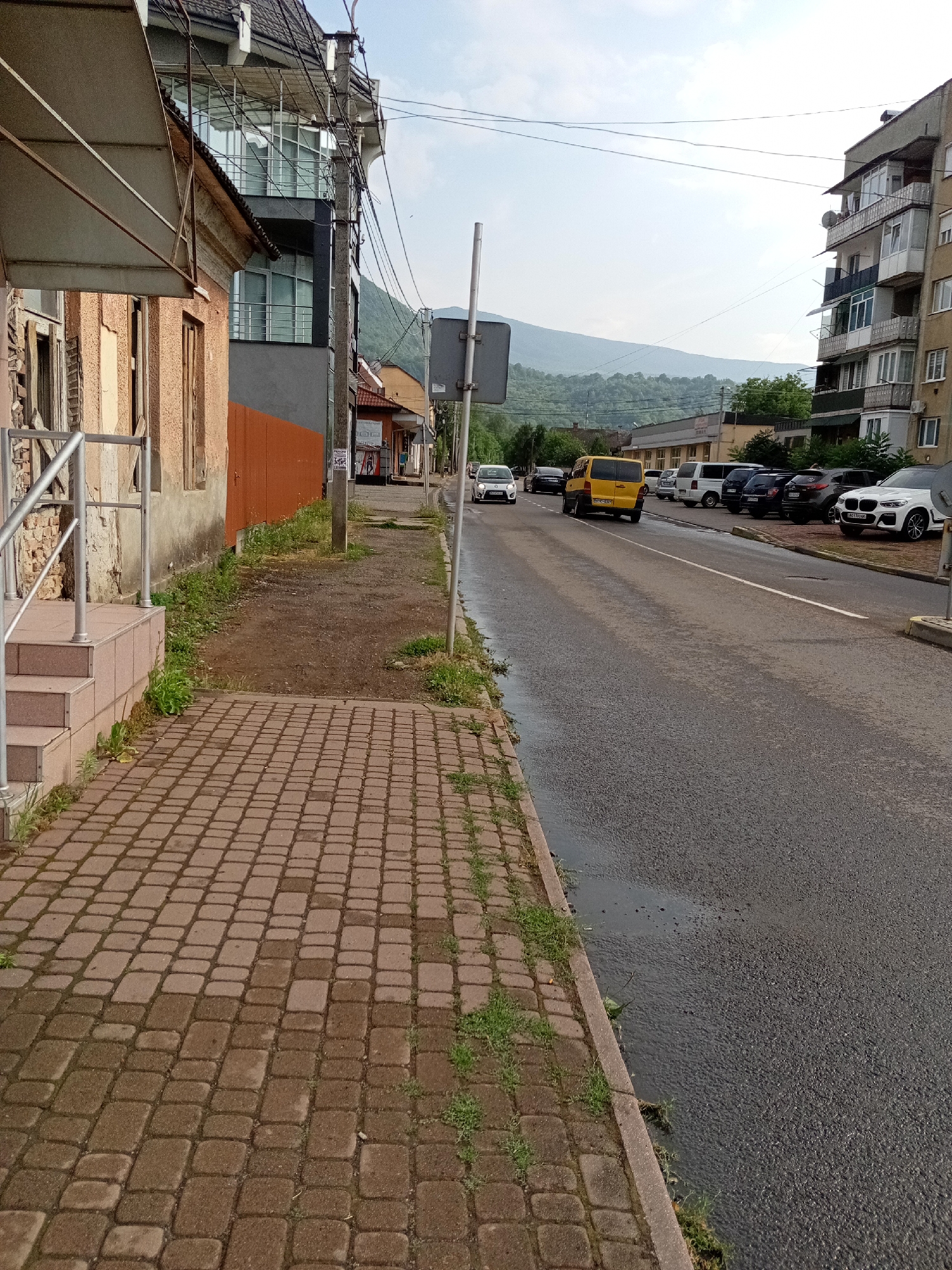 This is a street-level photograph taken in Tiachiv, Ukraine, likely during the daytime given the bright sky and shadows. The scene depicts a paved road with several vehicles parked along the right side and driving on it. A yellow minivan is in motion, followed by other cars, including a white BMW SUV parked near the foreground. The left side of the road features a sidewalk made of brown pavers, bordered by patchy grass and a weathered building with a light brown facade. Further down the street, more buildings are visible, including a multi-story apartment building with balconies and a row of shops. In the background, rolling hills covered in greenery suggest a mountainous or hilly landscape. The sky is clear and blue with a few wispy clouds. There are some puddles on the asphalt, possibly indicating recent rain. A street sign is visible on the right side of the road. No people are clearly discernible in the image. The overall impression is a somewhat ordinary street scene in a town with a natural, scenic backdrop.