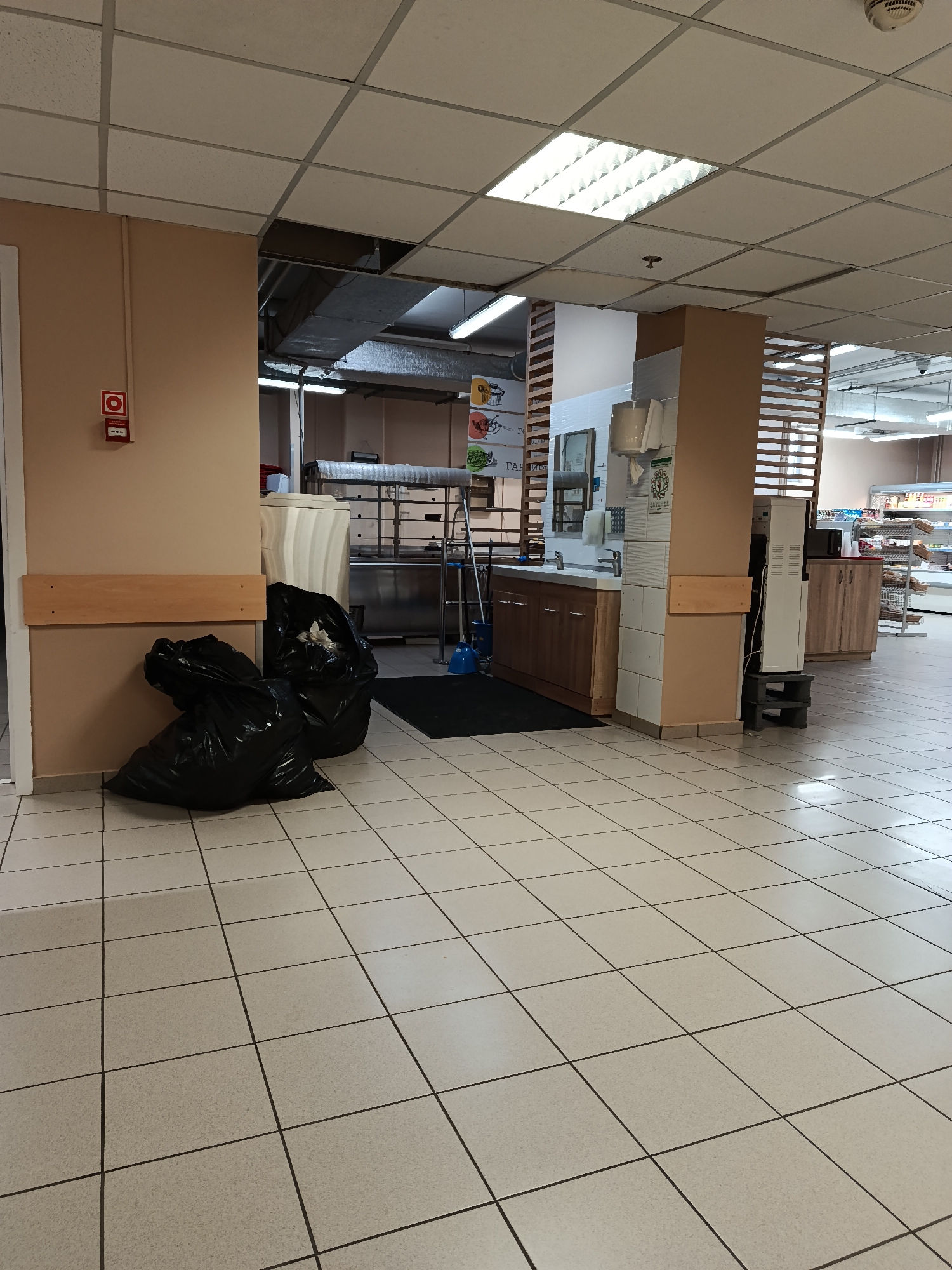 The image shows an interior view of a commercial space, likely a supermarket or food processing area. In the foreground, two large black garbage bags are positioned against a wall. The floor is covered in light-colored square tiles.
Through an opening in the wall, a secondary area can be seen. This area contains stainless steel equipment, likely for food preparation. A sink area with wooden cabinets is also visible, along with what appears to be a water purification unit and a display of some kind.
The lighting is provided by recessed fluorescent ceiling lights. The location is presumed to be Moscow, Russia, based on the prompt, but there are no visual elements within the image to independently confirm this. No people are visible.