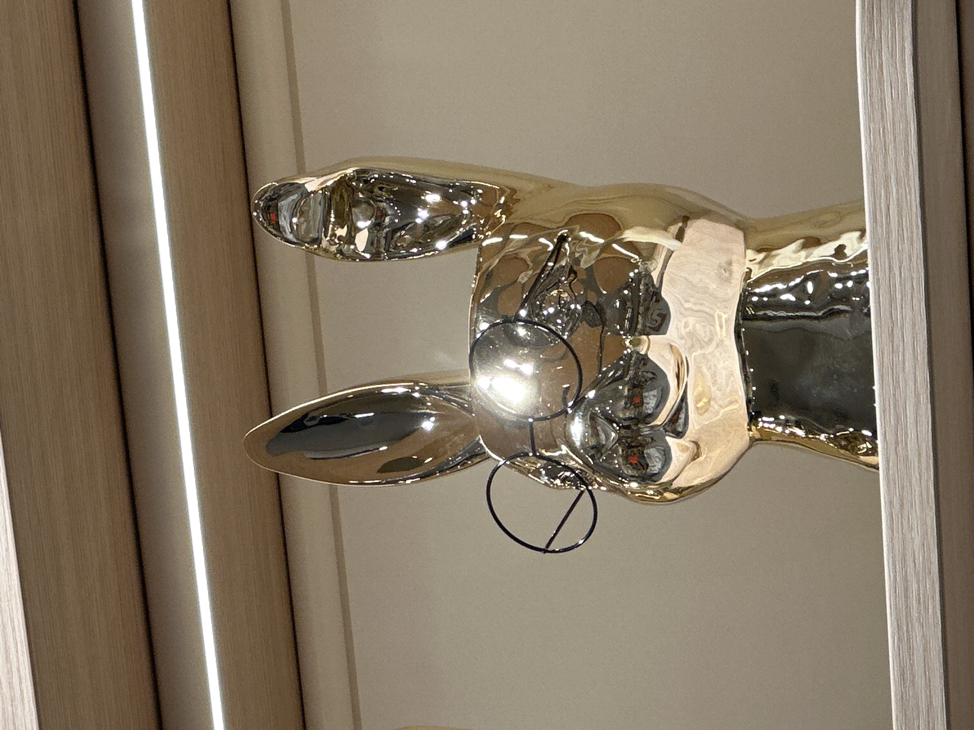 An abstract, highly reflective sculpture dominates the center of the image, appearing to be a stylized, elongated human arm or hand made of polished golden metal. The sculpture is oriented horizontally, with the "hand" or "finger" pointing towards the left side of the frame. Its surface is exceptionally shiny, creating numerous bright highlights and distorted reflections of the surrounding environment, including what appear to be overhead light sources and perhaps parts of the room. The material exhibits a somewhat rippled or undulating texture, which contributes to the organic, flowing appearance and the warped reflections.
Near the central part of the sculpture, where a palm would be on a human hand, there is a dark, thin, wire-like structure. This structure forms a distinct circle with a diagonal line crossing it, and it seems to be integrated with the sculpture itself, possibly holding a small, spherical element or a light source, as a bright glare emanates from this area.
The background consists of neutral-toned interior walls or ceiling, likely a light beige or off-white color. On the left side of the frame, a vertical wooden panel with a finely textured grain is visible. Running vertically alongside this panel is a bright, luminous strip of white light, suggesting an embedded LED fixture or similar architectural lighting. A similar vertical wooden element, possibly a door frame or another decorative panel, is present on the right side of the image, framing that edge of the composition. The entire scene is brightly lit by artificial light. No people or text are visible in the image.