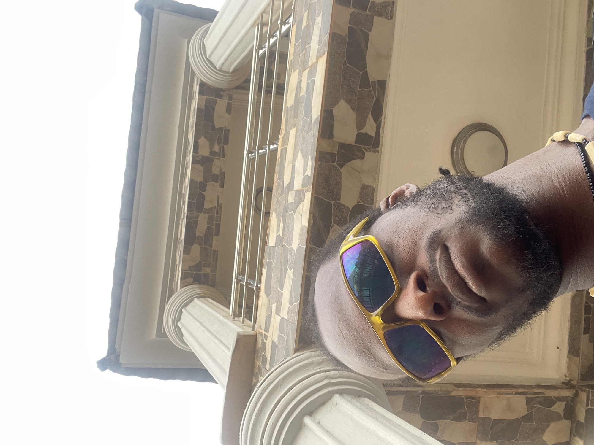 The image, taken from a low-angle perspective and rotated 90 degrees counter-clockwise, features a person looking downwards towards the camera. The subject is a male with dark skin and a full beard, wearing prominent yellow-framed sunglasses with reflective blue and purple lenses that obscure his eyes. A beaded bracelet is visible on his right wrist.
The background reveals architectural details of a building, characteristic of an outdoor setting like a porch or balcony. To the left and right, large white columns with classical fluting and decorative capitals are partially visible. The building's walls are adorned with textured, patterned tiles in varying shades of grey, brown, and off-white. Between the columns, vertical metal bars are present, likely part of a railing or security grill. Behind the person's head, a circular fixture, possibly a light or vent, is embedded in a plain, light-colored wall.
The upper left portion of the frame shows a bright, clear sky, indicating that the photograph was taken during the daytime. The overall impression suggests a casual portrait taken in an urban or residential environment in Abuja, Nigeria. There is no visible text in the image.