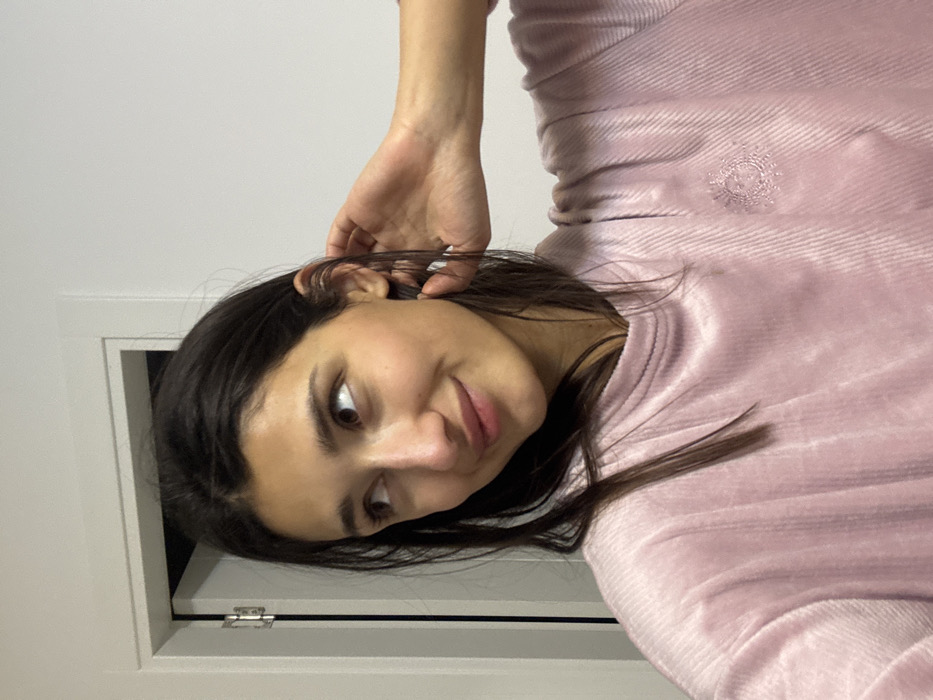 A young woman with dark hair tilts her head towards the camera, smiling subtly. She wears a soft pink, textured long-sleeved shirt. Her left hand is raised near her ear. The background features a plain white wall and what appears to be a partially visible door frame in the lower-left corner, with a metallic hinge. The lighting is soft and even, casting gentle shadows.