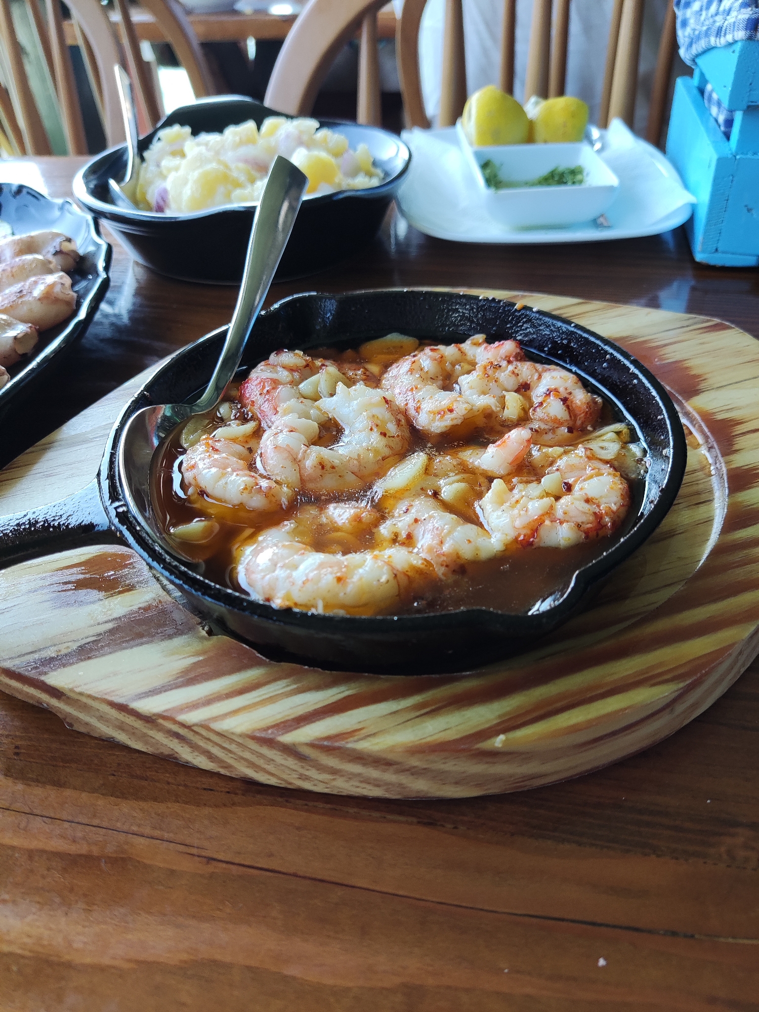 A close-up, high-angle shot shows a cast-iron skillet filled with succulent shrimp swimming in a savory broth, seasoned with red pepper flakes and garlic. The skillet rests on a wooden platter, which in turn sits on a wooden table. To the left, a small black plate contains several cooked prawns. In the background, a black bowl of potato salad is visible, accompanied by a spoon. Further back, a white plate holds two lemons and a small dish of green herbs, next to a bright blue wooden condiment holder. Chairs with wooden slats are visible in the blurred background, suggesting an indoor dining setting, likely in Gornji Štoj - Shtoji i Epërm, Montenegro.