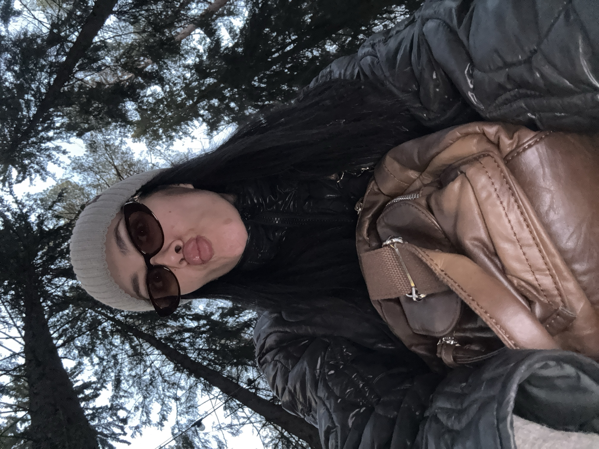A low-angle, close-up outdoor portrait features a young person with long dark hair, playfully sticking out their tongue and making a funny face towards the camera. They are wearing a light-colored knitted beanie and dark, thick-framed sunglasses. Their attire includes a black quilted jacket, and a brown leather-like shoulder bag with straps is visible on their left side.
The background consists of a dense canopy of dark tree branches, some with sparse foliage, set against a bright, overcast sky. This indicates a wooded or forested environment. The light is soft and diffused, typical of an overcast day, suggesting cool weather given the subject's winter attire. The person's expression conveys a sense of lightheartedness and humor. No text is visible in the image. The setting is a wooded area in Votkinsk, Russia.