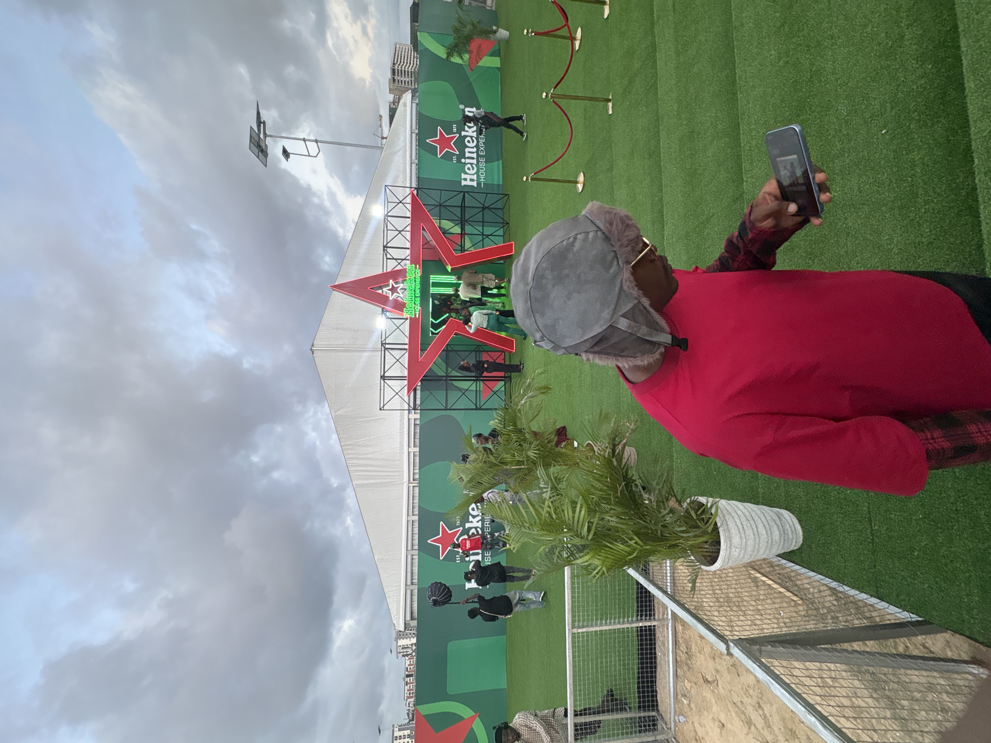 A medium shot, taken vertically, shows a man taking a picture with his phone. He is wearing a red shirt, a gray hat, and glasses. The background features a large outdoor Heineken event in Lagos, Nigeria. Behind him is a potted plant and a security fence with sand. In the distance is a Heineken sign and a large red star. People are visible entering the event, which is held on a green lawn under a cloudy sky.