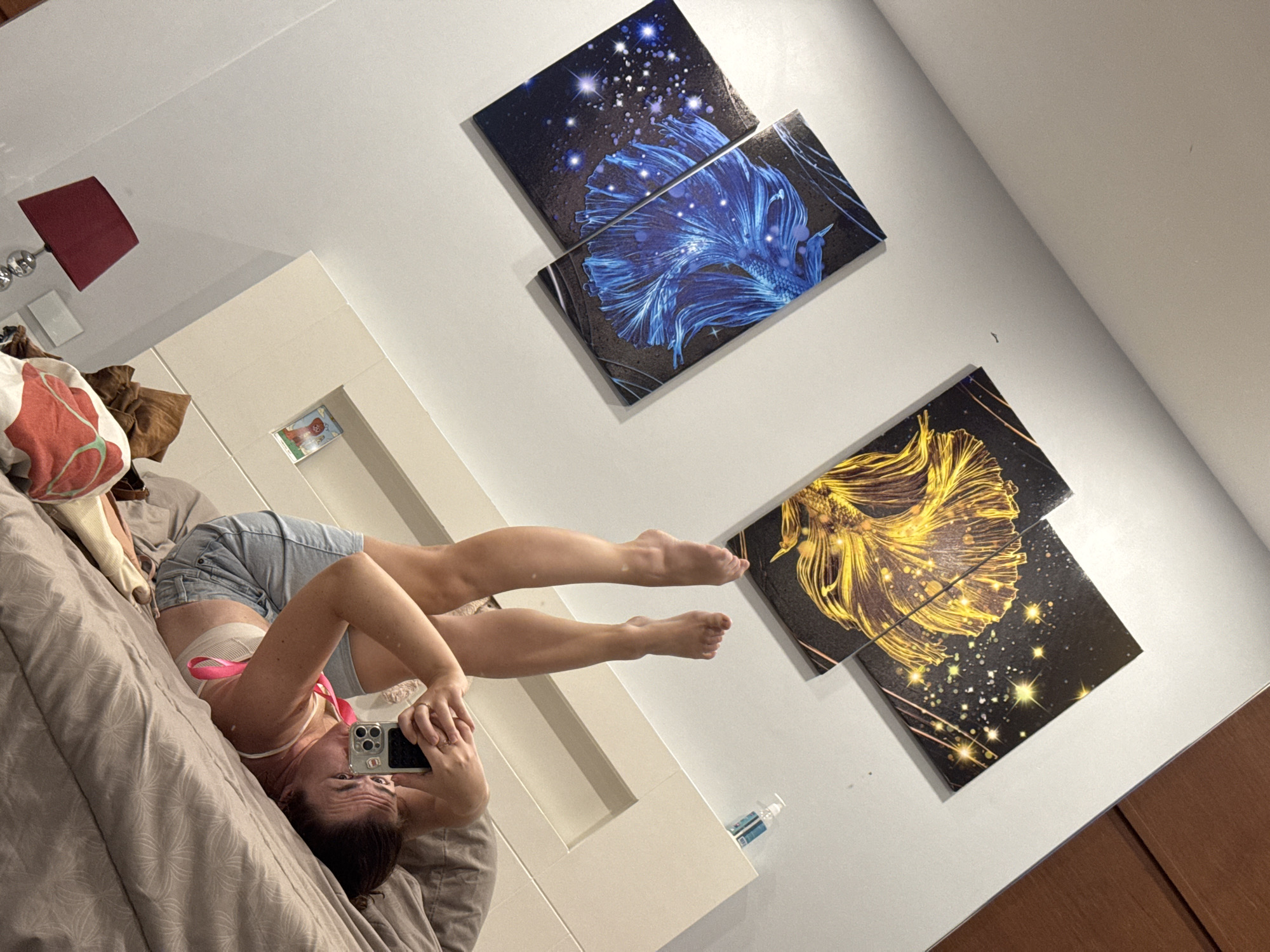 In Rawai, Thailand, a woman is lying on a bed, taking a selfie with her phone. Her legs are raised in the air, and she is wearing denim shorts and a bra. The room is decorated with two large paintings of betta fish, one in blue and the other in yellow, both set against a starry background. A bedside lamp with a red shade is visible, as is a hand sanitizer bottle on the headboard. The bed is covered with a textured grey duvet.