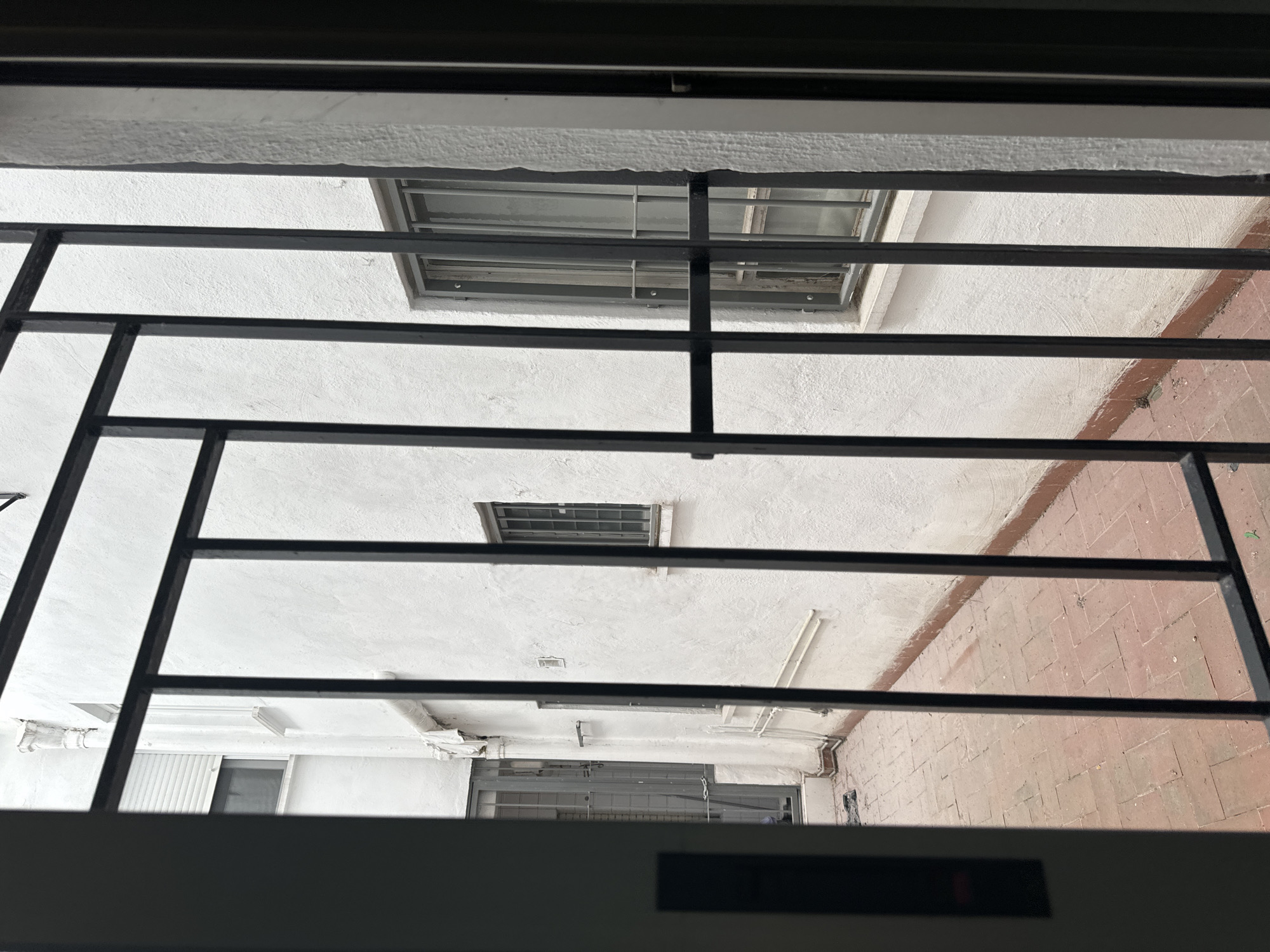 The image shows a view through a black metal gate into a narrow courtyard. The courtyard has a tiled floor and white stucco walls. There are several windows visible in the walls, some with metal security bars. On the left side of the courtyard, there is a metal door with a glass panel. The scene appears to be in an urban environment, possibly residential. The presence of metal bars on the windows and doors suggests a need for security. The lighting suggests it is daytime, but the sky is overcast, creating a diffused light. The overall impression is one of a functional, perhaps slightly utilitarian, outdoor space. The style of the architecture and the presence of terracotta tiles are common in southern European cities like Murcia, Spain.
