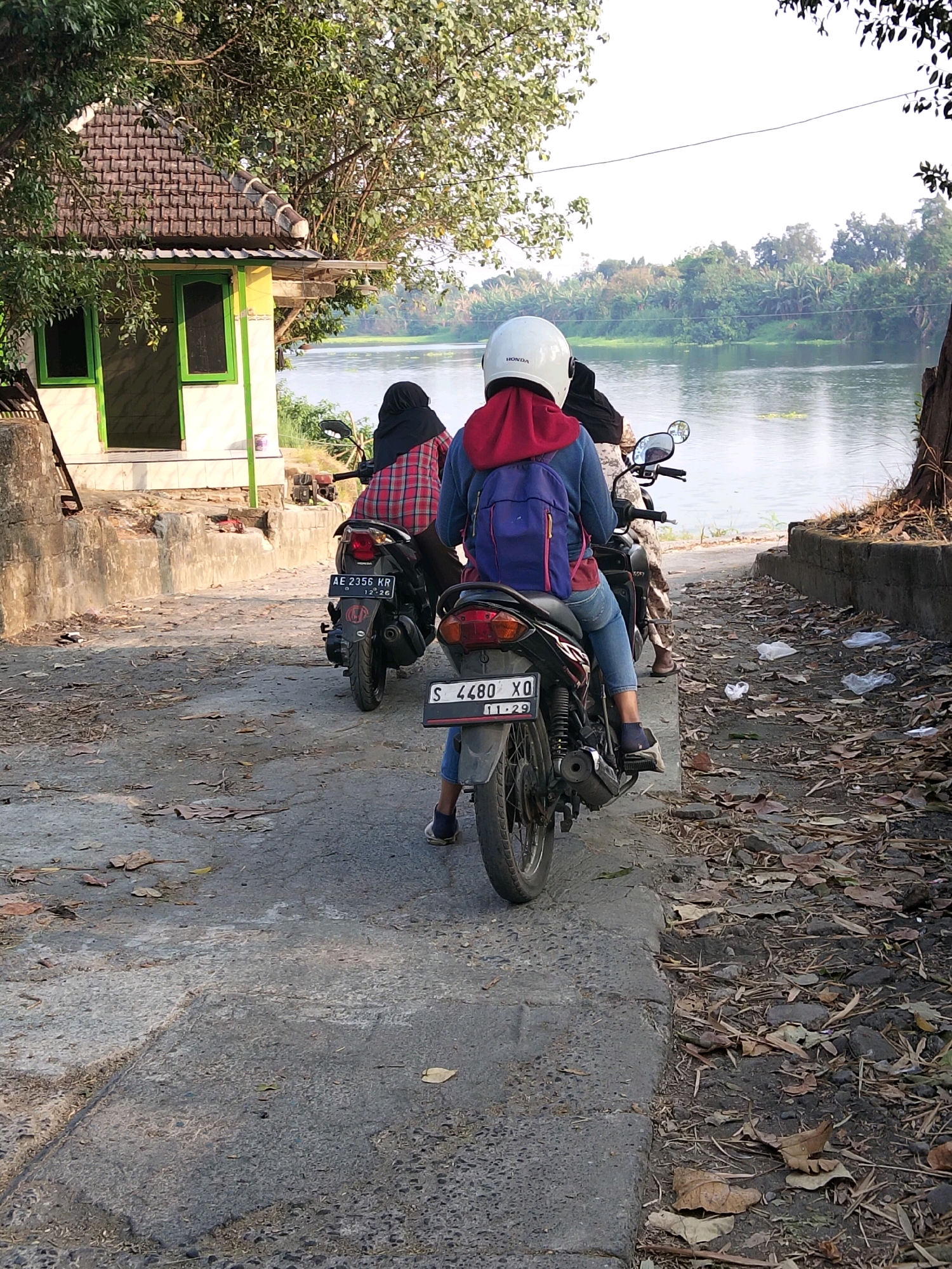 Two people on a motorcycle are parked on a paved road near a body of water. The person in the front is wearing a white helmet, a red hoodie, and blue jeans, with a blue backpack. The person behind them is wearing a black hijab and a red and black plaid shirt. The motorcycle has a license plate that reads "AE 2356 KR". In the background, there is a small building with a tiled roof and green-framed windows, trees, and a river. The ground is covered with leaves and debris. The scene is likely in Kesamben, Indonesia, given the license plate format.