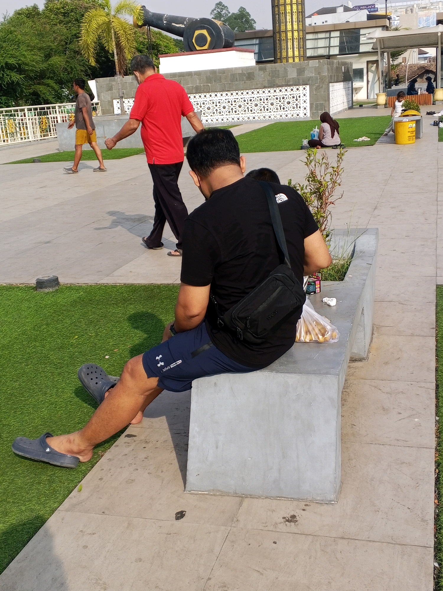 In an outdoor public space in Serang, Indonesia, a man wearing a black t-shirt, navy blue shorts, and grey Crocs sits on a concrete bench. A black crossbody bag is slung over his shoulder. To his left, a plastic bag filled with what appear to be snacks sits on the bench. Two other individuals are visible in the background: one man in a red shirt and black pants walks away from the camera, and another man in a brown t-shirt and yellow shorts walks to the left. A large cannon structure is visible in the upper left of the frame, with a palm tree and other foliage surrounding it. The ground is paved with concrete tiles, with sections of artificial green turf. A woman and child are seated on the grass in the mid-ground, near a yellow trash receptacle. The overall scene depicts a daytime outdoor gathering in an urban park setting.