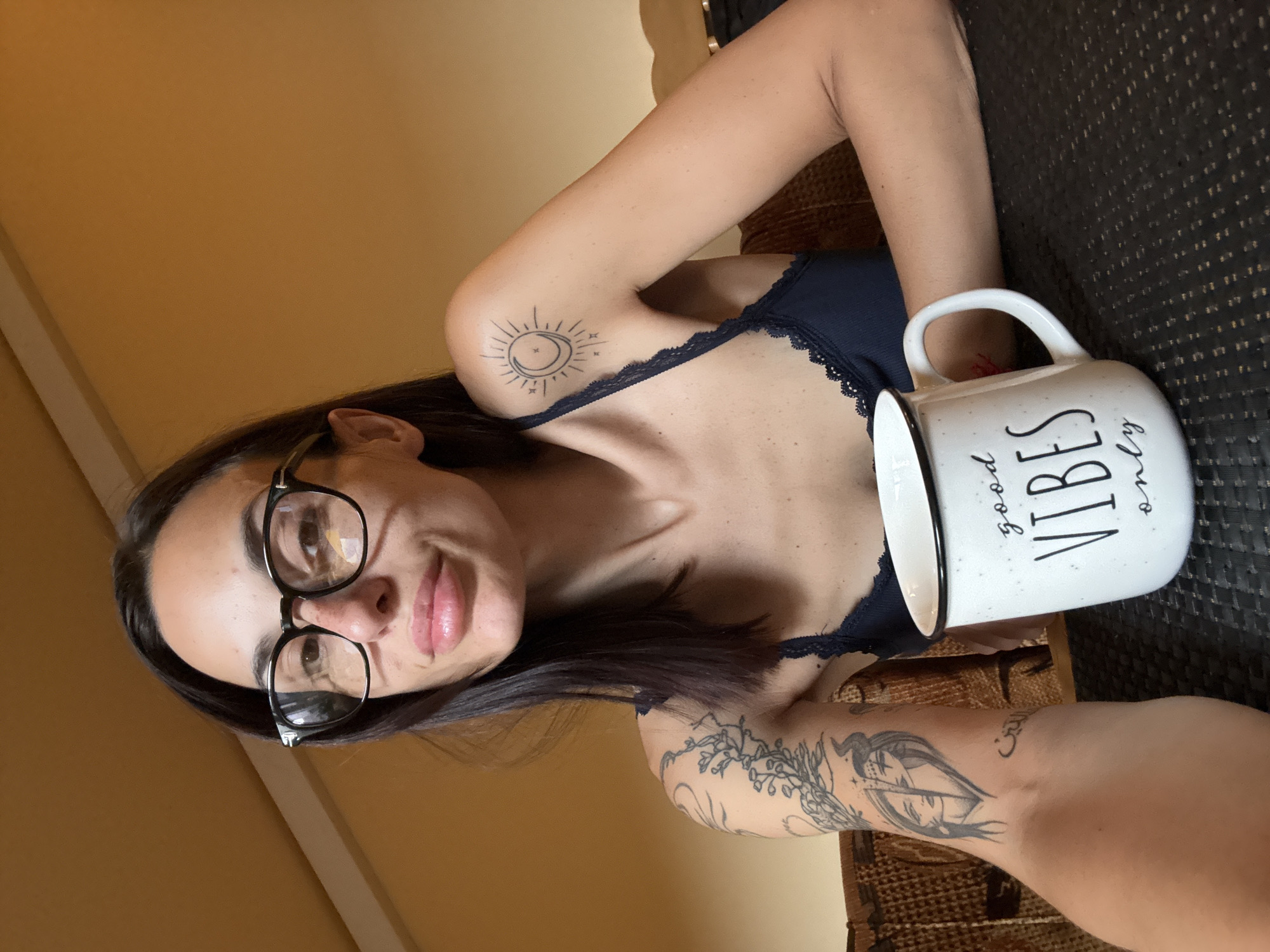 The image is a close-up, sideways selfie of a young woman taken indoors, presented with a warm, soft lighting that suggests daytime. The woman is reclining or lying down, looking directly at the camera with a slight smile. She has dark hair and wears black-framed eyeglasses. Her attire includes a dark-colored top with a delicate lace trim visible on the strap.
She prominently displays several tattoos. On her left shoulder/upper arm, there is a circular outline tattoo depicting a stylized sun and crescent moon design, surrounded by radiating lines and small stars. On her right forearm, a larger, more complex tattoo features an intricate design that includes what appears to be a stylized face or figure within an abstract form, possibly resembling an arrow or feather, intertwined with leafy or floral patterns. Faint Arabic script is also discernible within this forearm tattoo.
In her right hand, she holds a white ceramic mug with a black rim and handle. The mug faces the camera, displaying the phrase "good VIBES only" in black lettering. "Good" and "only" are written in a cursive font, while "VIBES" is in a bold, capitalized sans-serif font.
The background primarily features a solid, warm tan or ochre-colored wall. To the right, behind her arm and the mug, a dark, textured surface, possibly a woven material like wicker or rattan, is visible. The overall impression is one of relaxation and comfort. The image is contextualized as being from Pleven, Bulgaria.
