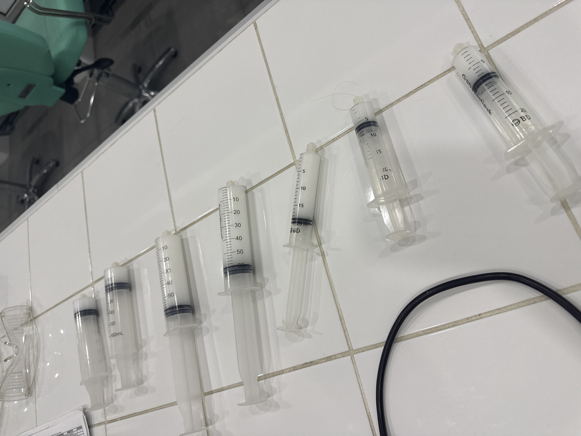 The image shows a collection of used syringes lying on a white tiled surface, possibly a countertop or floor. The syringes vary in size, ranging from smaller ones marked with volume measurements up to 20mL, to larger 60mL syringes. Markings such as "BD Plastipak" and "BD" are visible on some of the syringes. An unspecified black cable is visible in the bottom right corner. A green chair and its base can be seen in the background. There is no information in the image to indicate that it is located in the city of San Bernardino Tlaxcalancingo, Mexico.