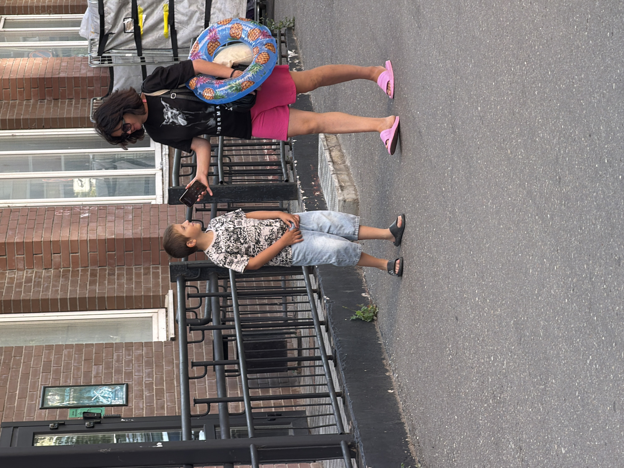 In an urban outdoor setting, a woman with dark, tousled hair and sunglasses stands holding a smartphone in her left hand, offering it towards a young boy. She is wearing a black t-shirt with a graphic of a dog, hot pink shorts, and pink slides. Around her right arm, she carries a blue inflatable swim ring patterned with pineapples. The boy, dressed in a grey and white patterned t-shirt and light blue denim shorts, stands facing the woman, looking at the phone. He is wearing dark slides. They are positioned on a paved asphalt surface in front of a brick building with a dark metal fence. The background includes windows of the building and some industrial-looking equipment on the right.