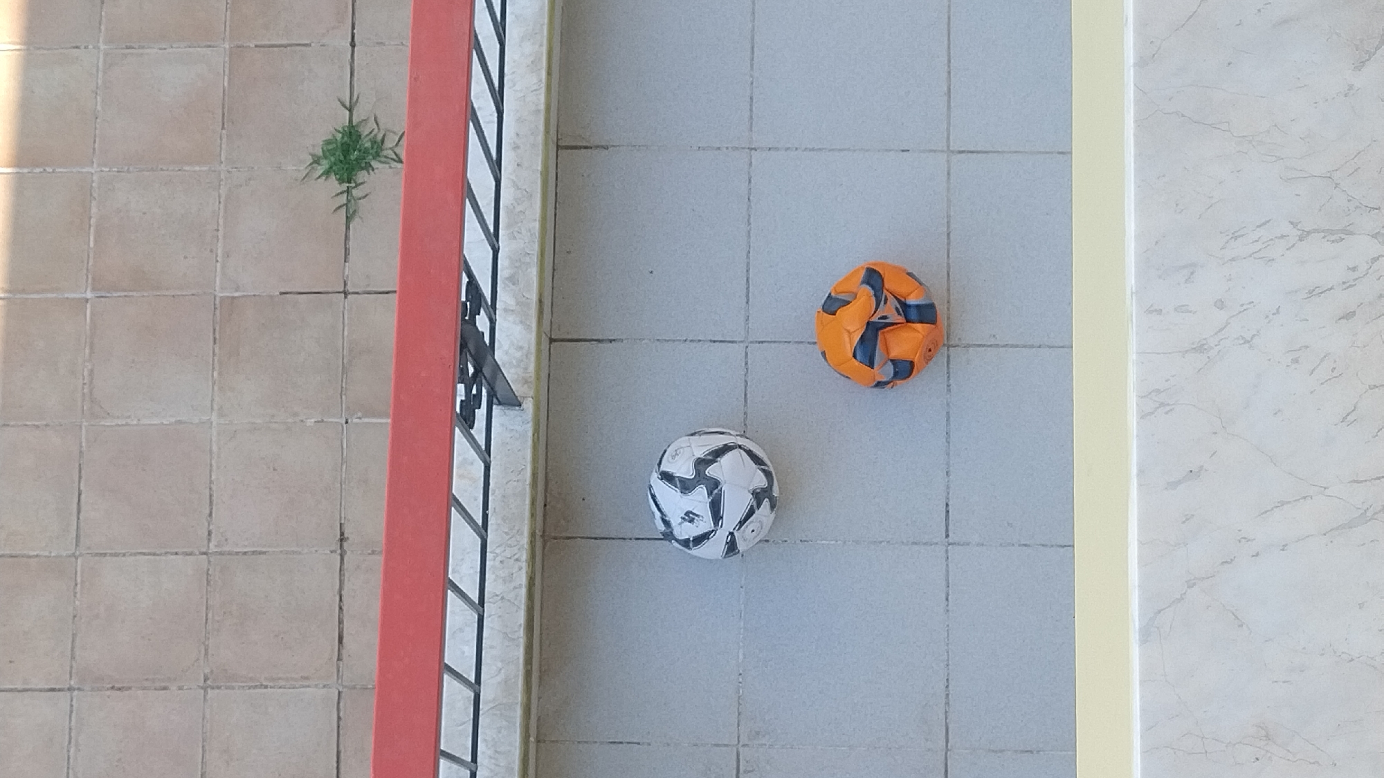 Two deflated soccer balls are on a tiled surface. One ball is white with black markings, and the other is orange with blue markings. A red metal railing is visible in the upper portion of the image, and a pale yellow ledge and marble surface are in the lower portion. A small plant grows from a crack between the tiles in the upper right corner. The scene appears to be outdoors, possibly on a balcony or patio. This image was taken in Perea, Greece.