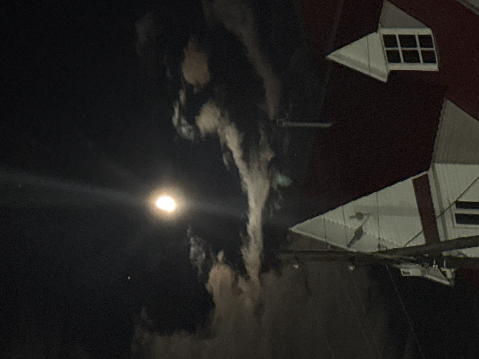 The image shows a night scene. In the upper right corner, there is a red building with white trim and windows. In the sky, a bright moon is visible with a prominent light flare. There are clouds in the sky near the building. There is a visible power line/cable running to the building. The location is identified as Прикамский, Russia.