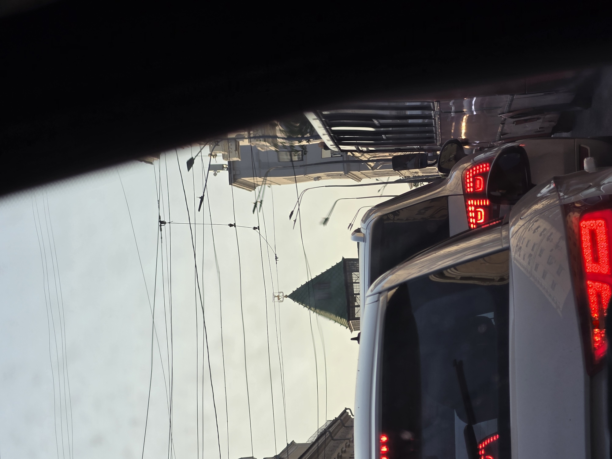 The image captures a view of traffic congestion on a city street in Nizhny Novgorod, Russia, likely in the historic center, given the architectural style of the buildings and the presence of a distinctive green-roofed tower. The perspective is from inside a vehicle, looking forward through the windshield and side windows, with a dark, blurred element, possibly a pillar or part of the vehicle's interior, dominating the right side of the frame.
In the foreground, the rear lights of several cars are prominently visible, indicating they are stopped or moving slowly. The red brake lights of a light-colored car are sharp and in focus, showing a modern design. Behind it, another car's taillights are also illuminated. Further ahead, a bus is visible in the traffic lane.
The middle ground of the image features a distinctive green, pointed tower, likely part of an old building or church, topped with a weather vane that appears to be a standing animal, possibly a deer or a horse. The tower is surrounded by numerous overhead power lines, typical of urban Russian infrastructure. Adjacent to the tower, on the left, is a building with classical architectural elements, including arched windows and decorative cornices. On the right side of the tower, another multi-story building is visible, with a row of streetlights lining the pavement in front of it.
The sky is overcast with a pale, diffused light, suggesting it might be dawn, dusk, or a heavily clouded day. The overall scene conveys a sense of urban life and the common experience of traffic delays in a city with historical architecture.
