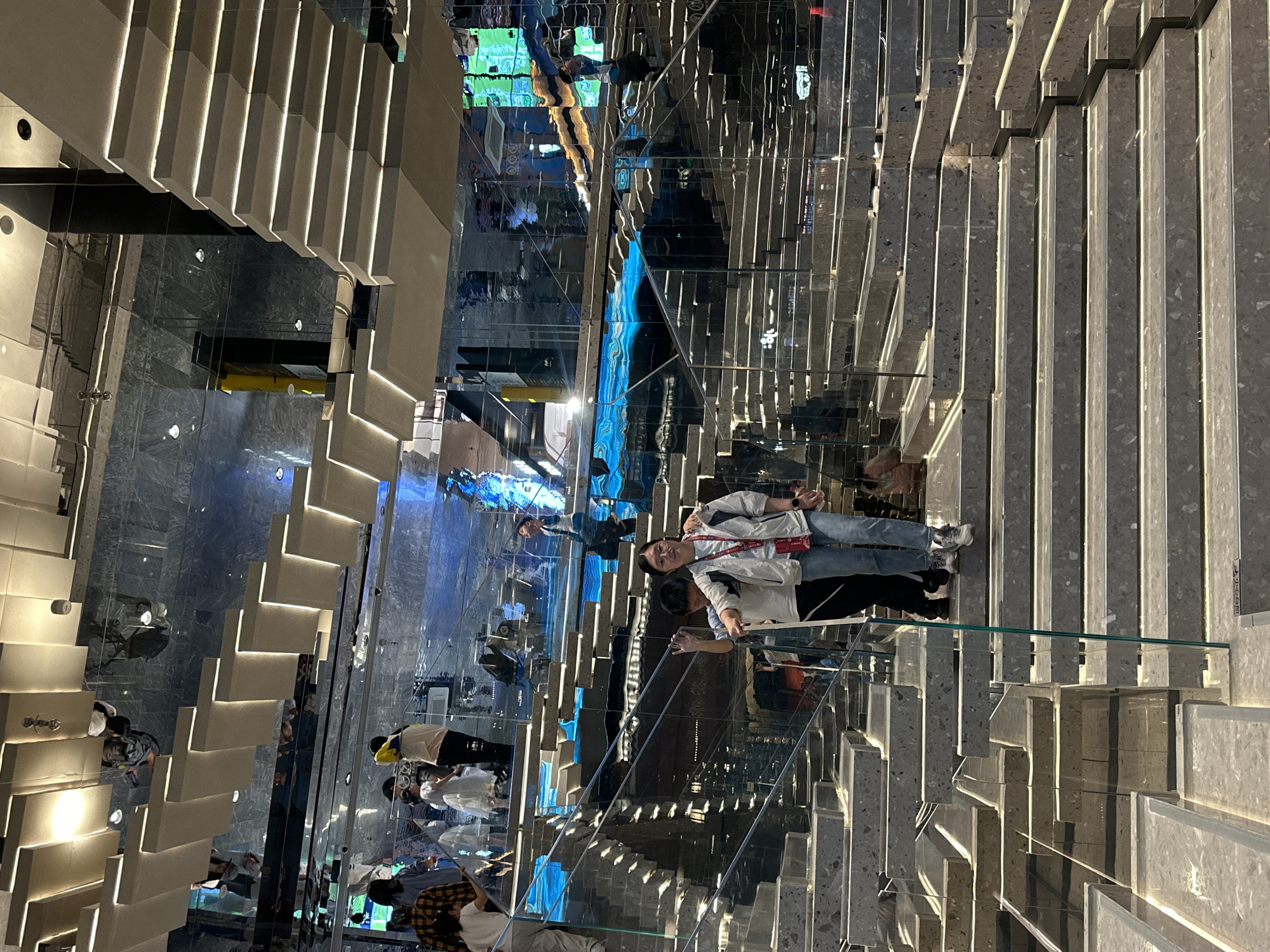 The image depicts an interior scene in a location that features staircases and reflective surfaces. A person is standing in the center of the image wearing a white jacket and blue jeans, smiling slightly. A second person is standing slightly behind the first, partially obscured. The surroundings consist of a complex arrangement of stairs with built-in lighting, creating a symmetrical and multi-layered perspective due to the mirrored walls and surfaces. The composition is highly structured with geometric forms of the steps and reflected patterns. Other people are visible in the background, either climbing the stairs or standing in the mirrored reflections. The general lighting is warm and bright. Based on the provided information, it is likely located in Yuzhong District, China.