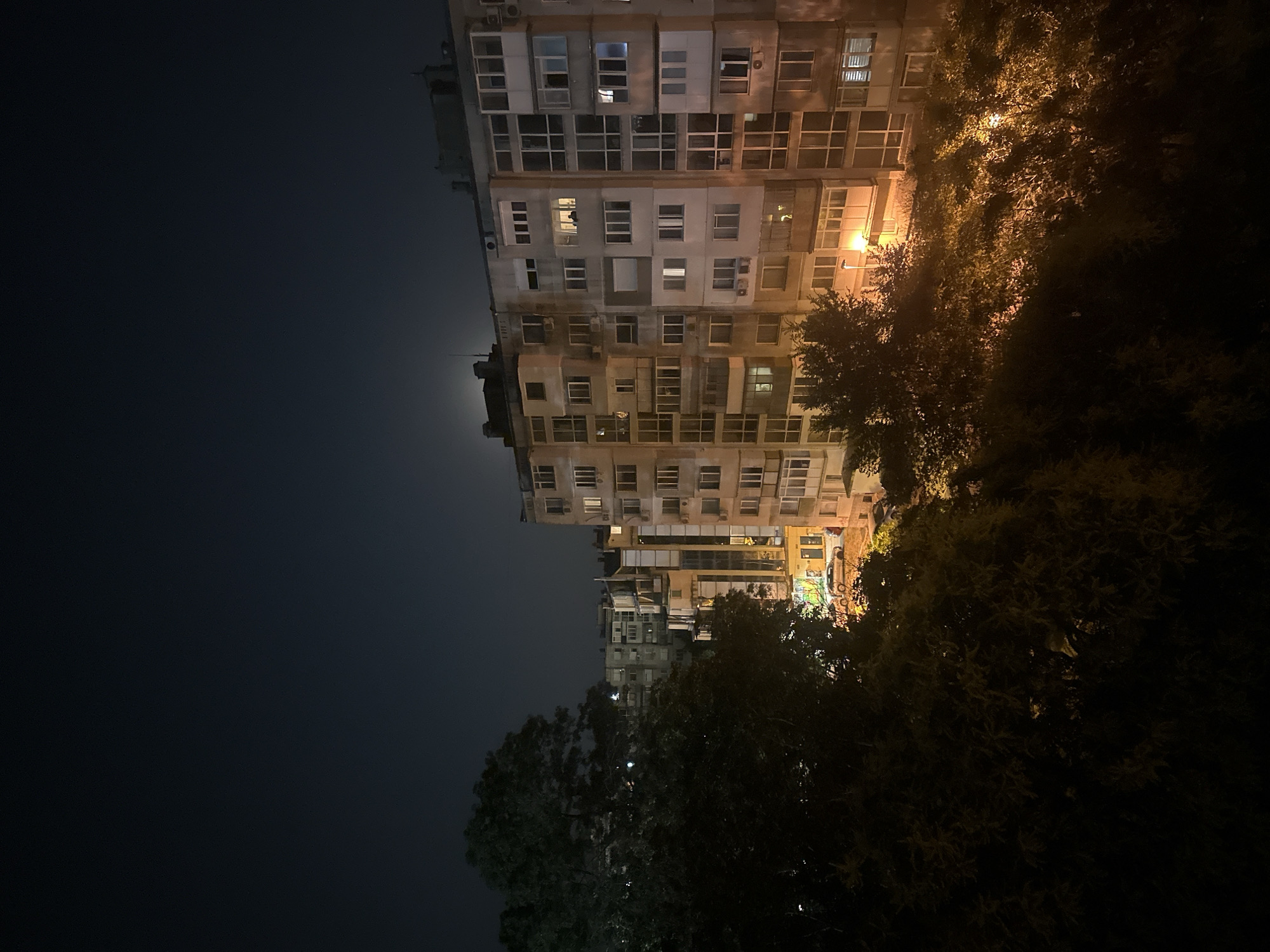 An aerial shot captures an apartment building at night in an urban setting. The sky is dark, with a faint moon visible above the building. Trees in the foreground are silhouetted against the illuminated windows of the building and the streetlights below. The building itself is multi-story with many windows, some of which are lit, suggesting occupancy. Cars are parked on the street in front of the building.