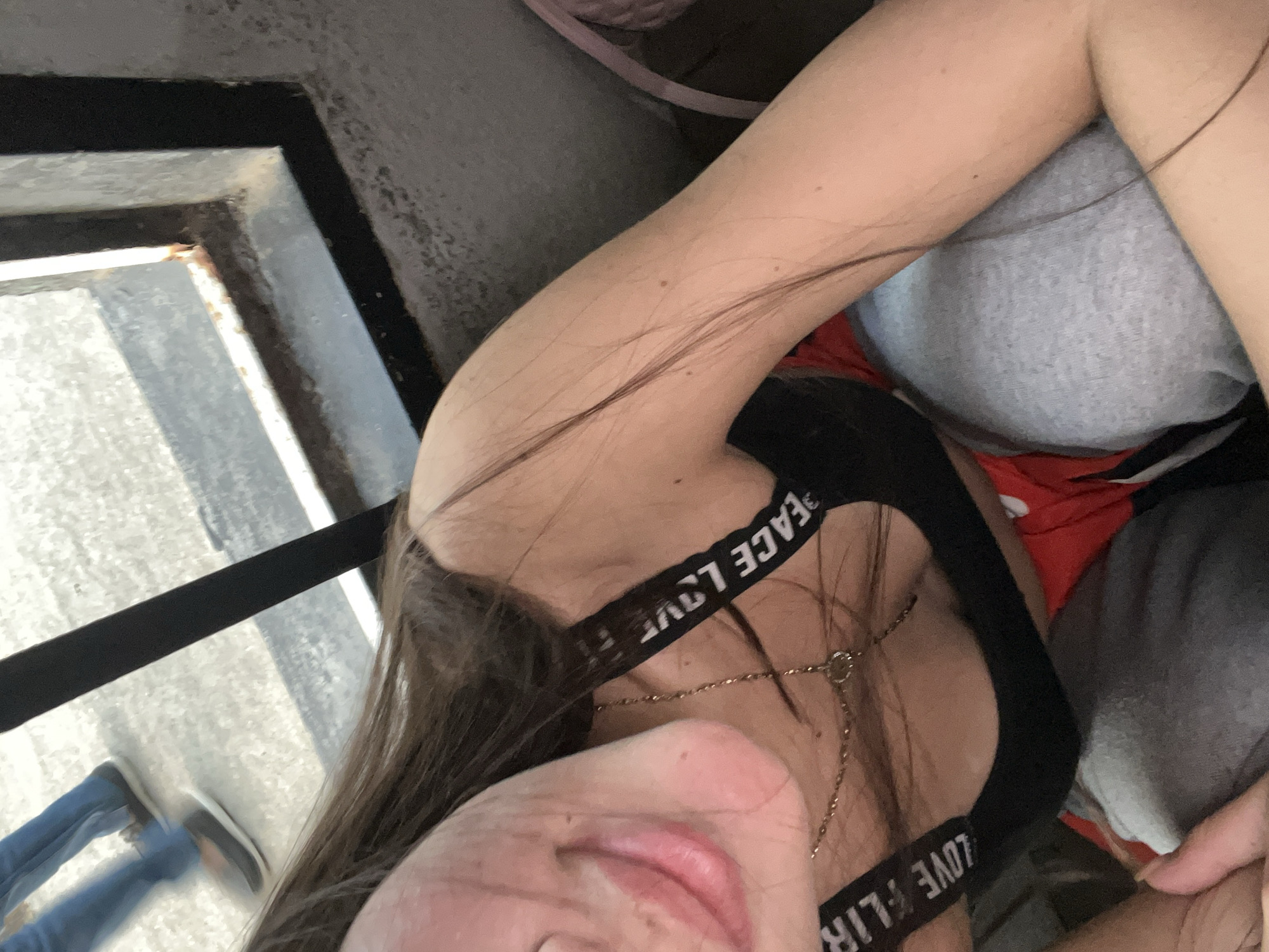 The image, taken in Juan Griego, Venezuela, presents a close-up, high-angle perspective of a person, likely female, in a relaxed setting. Her face, with pink lips and fair skin, is partially visible, looking towards the camera with a neutral or slightly smiling expression. Long, dark hair cascades across her shoulder and chest. She is wearing a black top or bra, with white text clearly visible on the strap, which reads "PEACE LOVE FLIRT". A delicate gold chain necklace with a small circular charm is worn around her neck.
The subject appears to be seated or reclining, with parts of her body resting against or covered by grey and red fabrics. In the background, through a dark, rectangular opening or frame (possibly a window or railing), an outdoor scene is visible. Bright natural daylight illuminates the setting, suggesting it is daytime. The legs of another individual, dressed in blue jeans and sneakers, are visible walking on a light-colored, paved surface beyond the frame. The overall impression is a candid, casual moment.