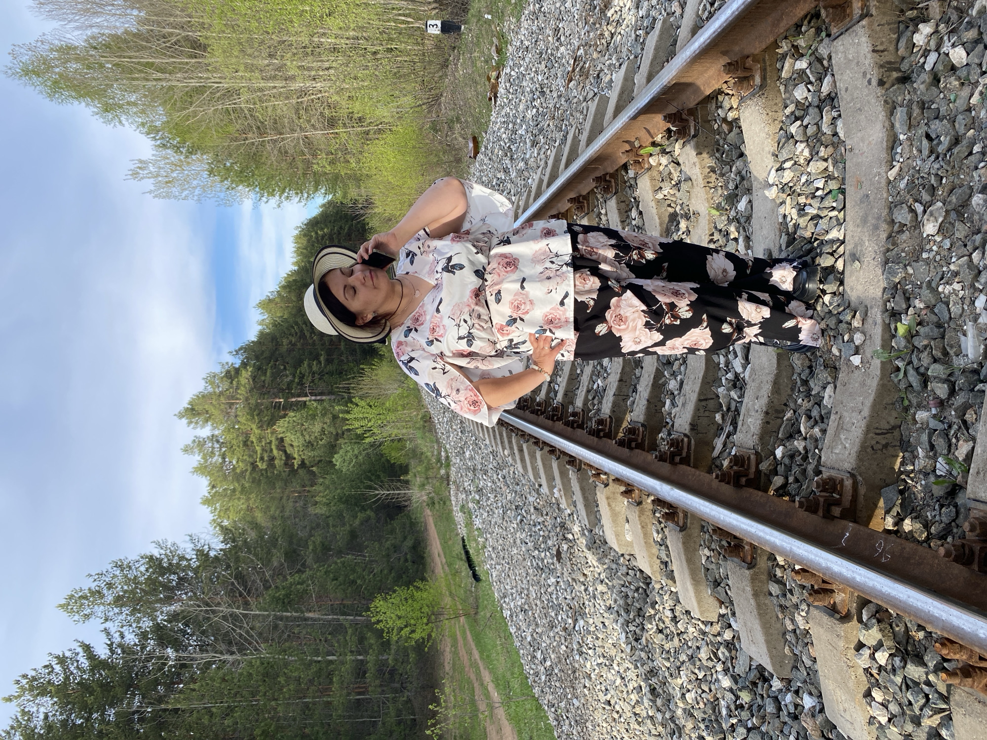 A woman stands on a railroad track in a wooded area, appearing to be near Chaikovsky, Russia. She is wearing a floral-patterned blouse, black pants with floral designs, and a hat. She is holding a cell phone to her ear, seemingly in conversation. The railroad track is surrounded by gravel and greenery. A small signpost with the number "3" is visible in the background. The day is sunny with a partly cloudy sky.