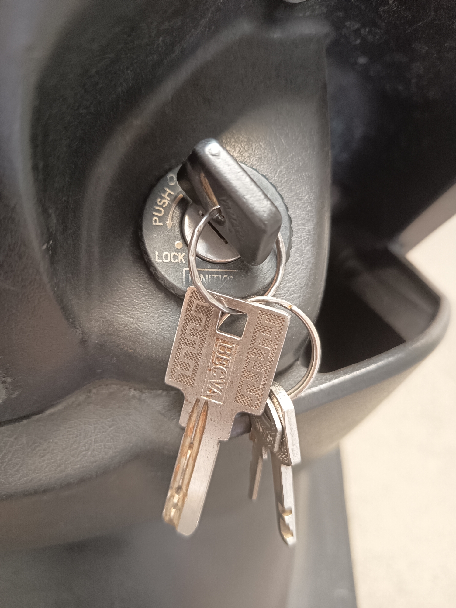 The image shows a close-up of a black ignition switch on what appears to be a scooter or motorcycle. A key is inserted into the ignition. Two other keys are attached to the same keyring and hang in front of the ignition. The keys are made of metal and have intricate patterns. The ignition switch has markings that read "PUSH," "LOCK," and "INT." The background is dark and out of focus, with some textured surfaces visible. The overall scene suggests a functional ignition system, possibly in a parked vehicle.