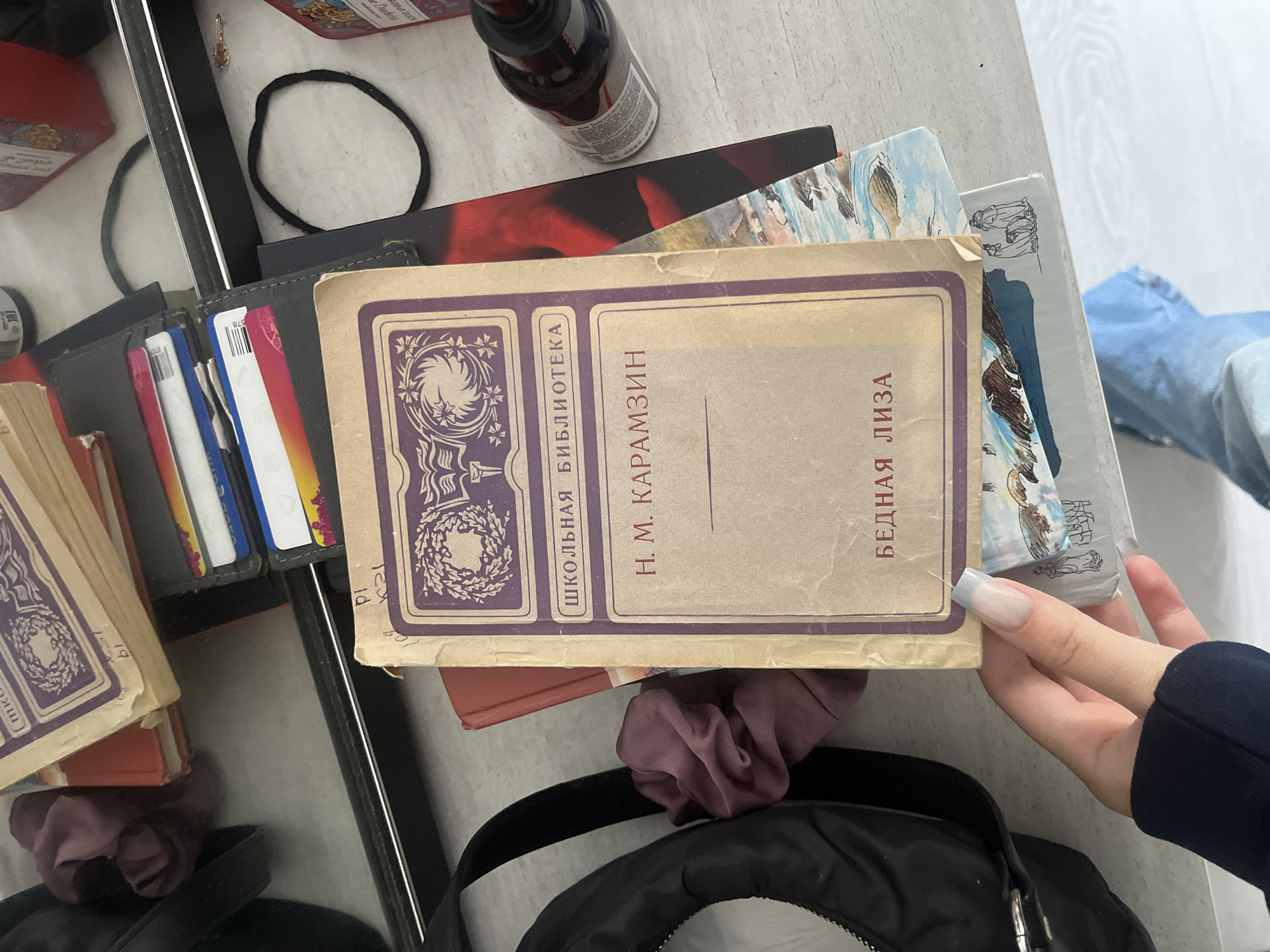 The image depicts a flat lay of various items on a white surface, possibly a desk or table. In the foreground, a person's hand with light gray nail polish is holding a book with a light brown cover featuring purple decorative elements and text. The title "Н. М. КАРАМЗИН" is visible, along with "ШКОЛЬНАЯ БИБЛИОТЕКА" and "БЕДНАЯ ЛИЗА".
Behind this book, other items are visible: a stack of books with a red cover, a black wallet containing several credit cards, and a dark-colored bag with a strap. A dark scrunchie or hair tie is placed to the left of the book in focus. Further back, a dark bottle with a dropper is present, and parts of other books or packaging are visible, including one with a red and black cover and another with a scenic illustration. The overall impression is of personal belongings scattered on a surface.
There is no indication of the city of Perm, Russia, or any specific urban location within the image. The scene is indoors, with the lighting suggesting an ambient environment rather than direct sunlight.