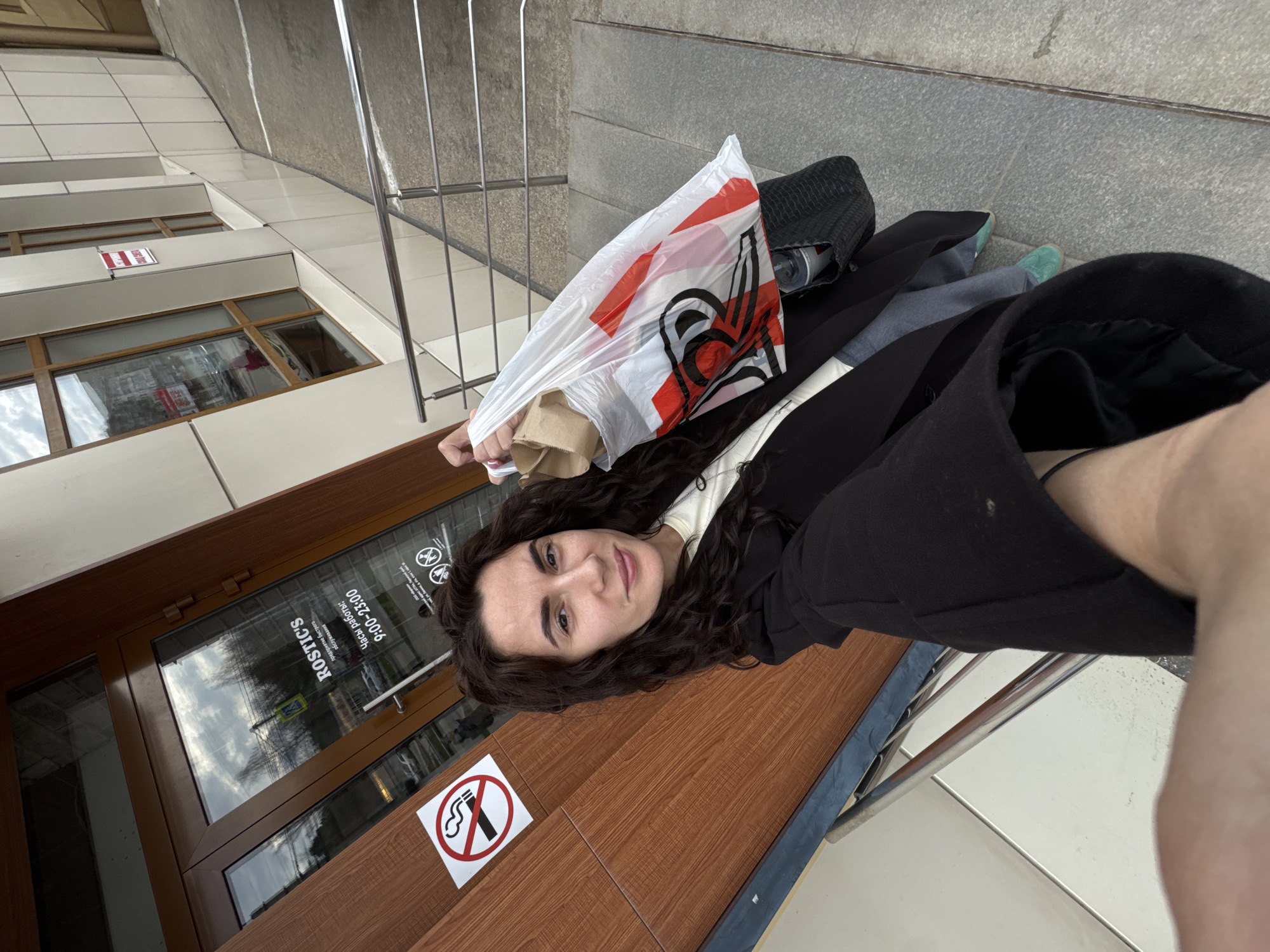 This image captures a selfie of a woman outdoors in Chaikovsky, Russia, taken from a tilted perspective. The woman, with dark, curly hair, has a calm expression with a slight smile, looking directly at the camera. She is wearing a dark jacket over a lighter top, light-colored pants, and noticeable turquoise/teal shoes.
She is holding a white plastic shopping bag with a prominent red and black logo, which resembles a stylized chicken or rooster head. Beside her, on the ground or a ledge, rests a black bag or purse.
The background shows the entrance of a commercial building with large glass doors and windows set in dark brown frames. Reflections in the glass indicate an overcast or partly cloudy daytime sky. Several pieces of text are visible: "ROSTIC'S" (reflected and inverted on the glass), "ЧАСЫ РАБОТЫ: 9:00-23:00" (Hours of operation: 9:00 AM - 11:00 PM), and smaller text below that mentions "доставка" (delivery) and "забираем быстро" (pick up quickly). A prominent "No Smoking" sign is affixed to a wooden surface next to the woman. A small red and white sign on the building's wall above the windows also reads "ВХОД" (Entrance). A metal railing is visible to the right, and the ground consists of light-colored tiled or concrete surfaces. The woman appears to be resting or waiting outside the establishment.