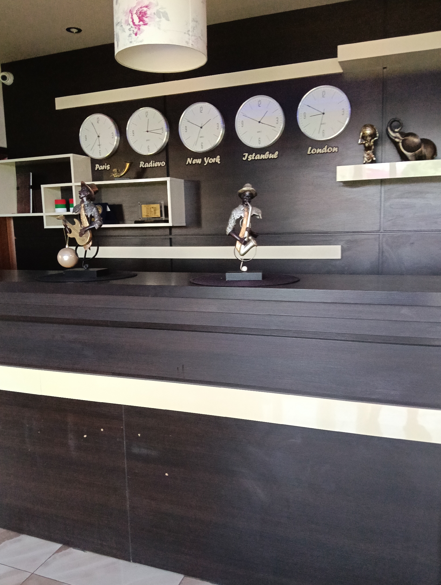 The image displays a modern interior scene, possibly a reception area, featuring a dark wood reception desk in the foreground. Above the desk, on a dark wall, are five world clocks displaying times for Paris, Radievo, New York, Istanbul, and London. Each clock is round with white faces and black hands. Below the clocks, on a white shelf, are decorative items, including a statue of a person playing a guitar and another statue of a person playing a saxophone. To the right of the statues, on the same shelf, is a bronze elephant figurine and a golden trophy or award. The overall ambiance suggests a sophisticated and organized space. The location in Radievo, Bulgaria, is indicated by one of the clocks.