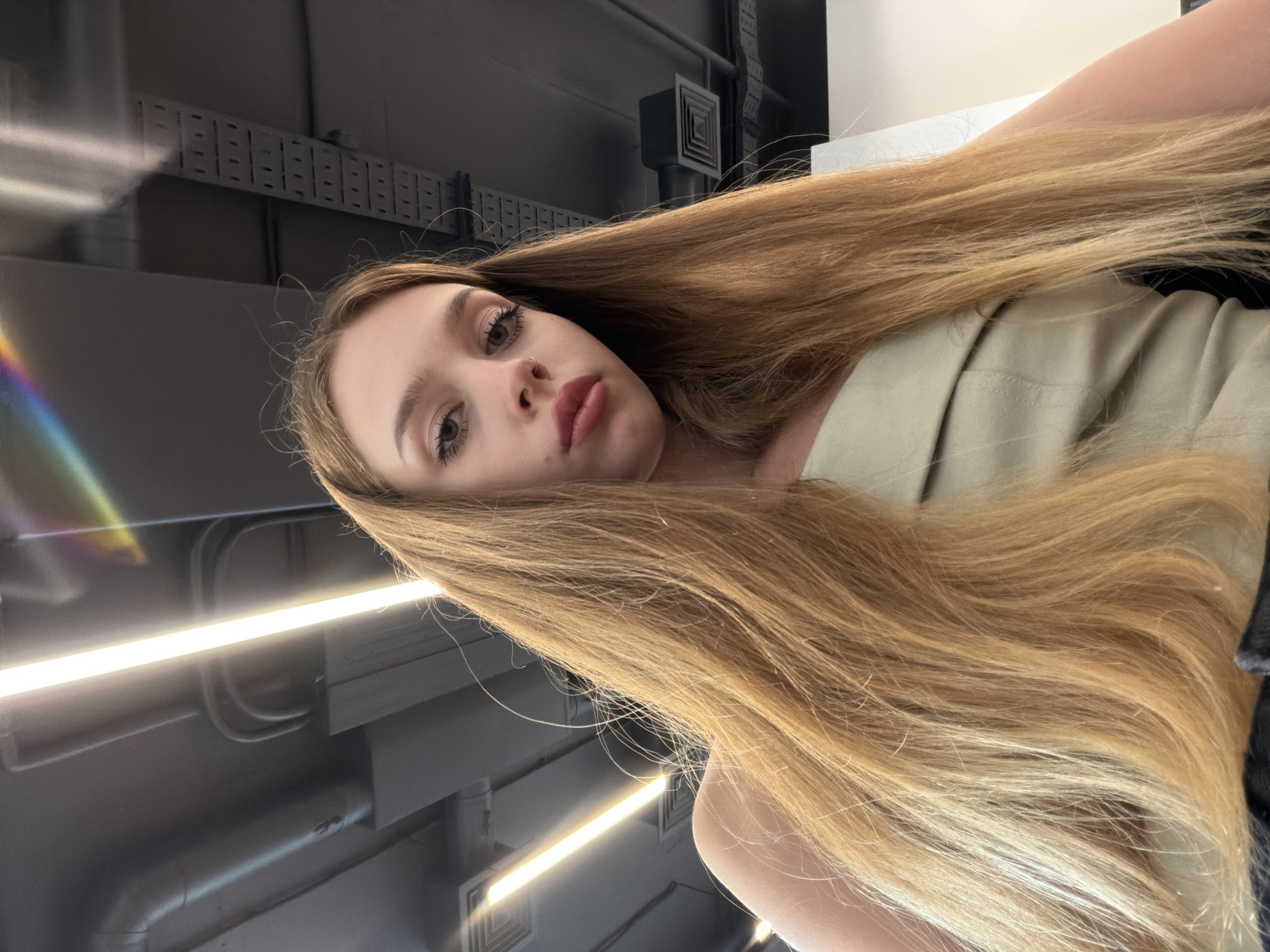 A young woman with long, blonde hair and fair skin is shown from a low-angle perspective, looking upwards towards the camera. She is wearing a light green top. Her makeup is subtle, with mascara and eyeliner visible. Her lips are slightly parted, and she has a small nose ring. The background is a ceiling with exposed pipes, ventilation systems, and bright fluorescent lights, suggesting an indoor commercial or industrial setting. There are rainbow-colored light refractions visible in the upper right corner of the image. The overall lighting is bright, with the fluorescent lights casting a strong glow.