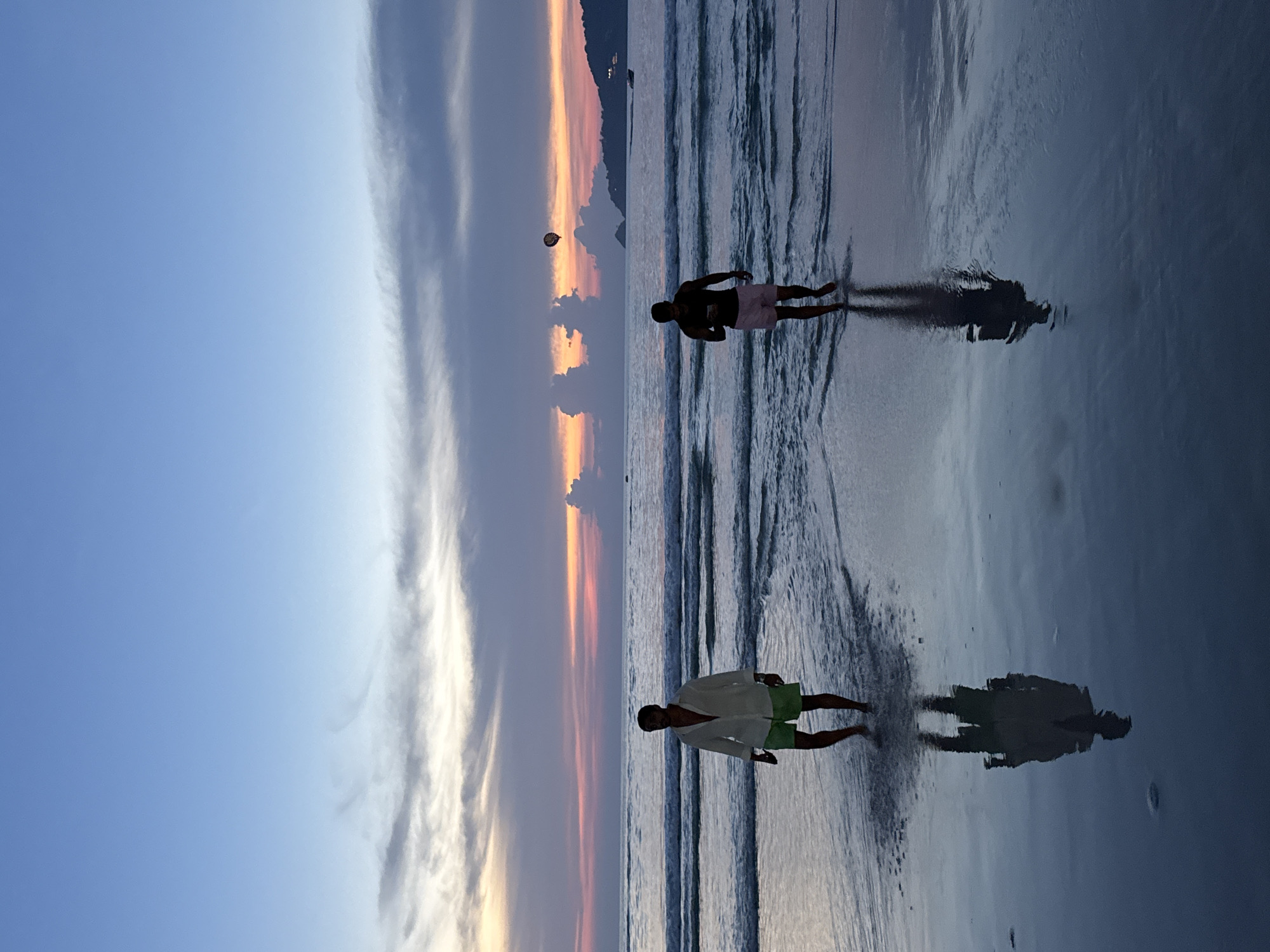 This is a vertical, full-shot photograph captured at eye level on a beach in Patong, Thailand, during twilight. The scene depicts two men walking in the shallow water near the shore. The sky is a gradient of colors, transitioning from a pale blue at the top to hues of pink, orange, and purple near the horizon where the sun has recently set. Wispy clouds streak across the sky, catching the fading light. The ocean is calm with gentle waves rolling in. The wet sand reflects the sky and the figures of the men.
The man on the left is wearing a white, open-collared shirt and light green shorts. He is walking towards the viewer. His reflection is clearly visible in the water. The man on the right is wearing a dark t-shirt and pink shorts. He is also walking towards the viewer, holding a phone in his right hand. His reflection is also visible. In the distance, a parasail can be seen in the air, and a boat is visible on the water. The shoreline curves into the distance, with a treeline visible on a distant hill. The overall impression is one of a peaceful evening on a tropical beach.