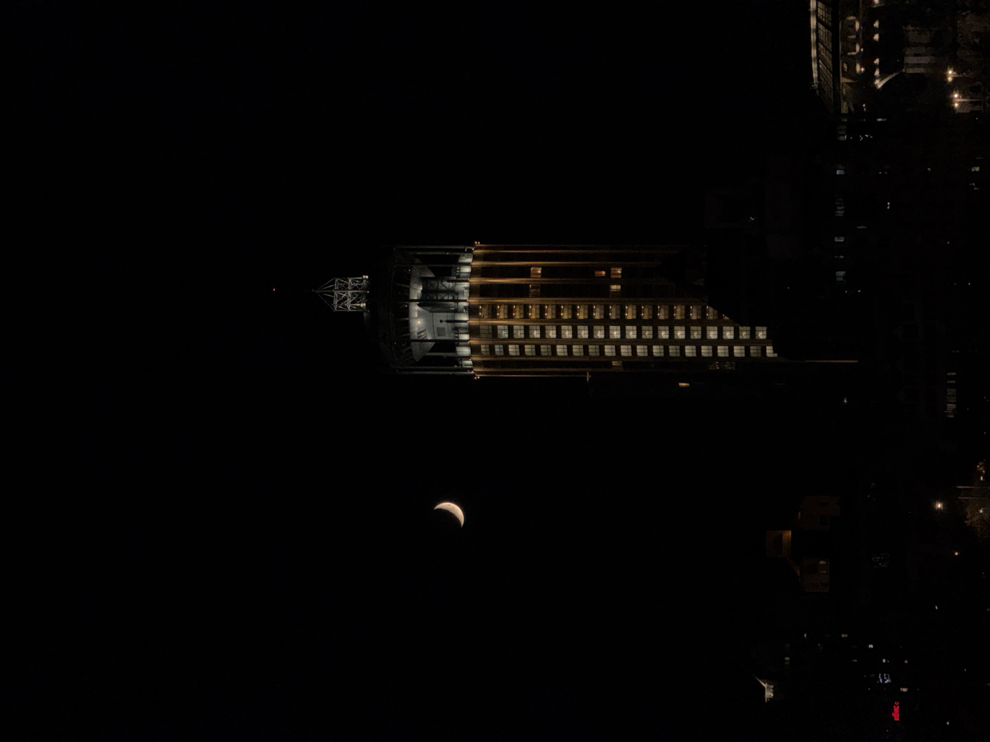The image captures a serene nighttime view in Moscow, Russia, dominated by a long, illuminated building stretching horizontally across the middle of the frame against a dark sky. The building, likely a hotel or office block, features numerous windows, many emitting a warm, yellowish light that forms a distinct grid pattern. On the far left of the building, a complex, lighter-toned structural element, possibly a part of the roof or an antenna, is visible.
Below the illuminated building and slightly to its left, a distinct crescent moon is visible. It glows with a reddish-orange hue, suggesting it might be close to the horizon or observed during a partial lunar eclipse. To the right of the main building and extending into the lower-right corner, faint scattered lights indicate other structures, streets, or possibly reflections from a body of water in the distant city. A red sign with indistinct, glowing Cyrillic text can be faintly seen in the bottom right corner. The overall scene is dark, emphasizing the artificial lights against the profound blackness of the night sky. No people or specific events are depicted, conveying a sense of quiet urban stillness.