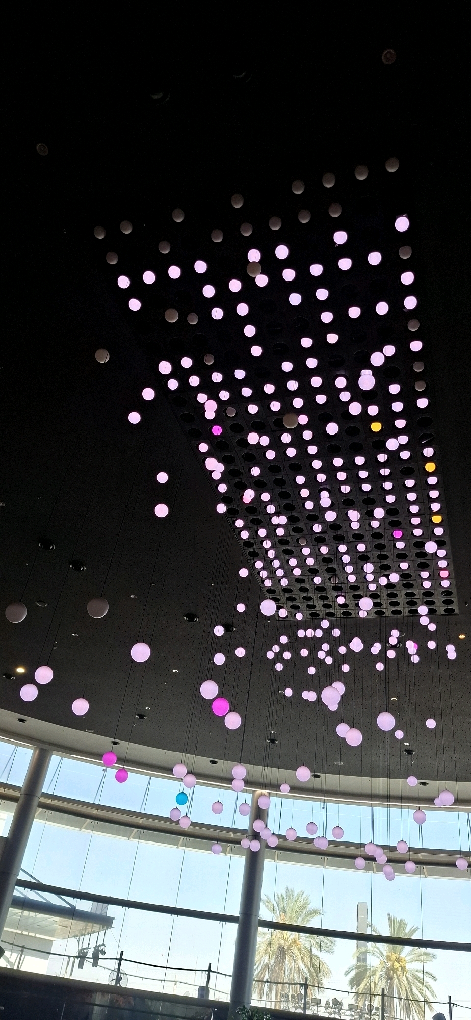 The image depicts a modern interior space with a black ceiling adorned with numerous spherical, illuminated lights. These lights are predominantly pink, with a few yellow and one blue orb. They are suspended at varying heights by thin black cords, creating a visually striking effect. Below the ceiling, a large, curved glass facade reveals an outdoor environment with palm trees and a bright blue sky, suggesting a commercial or public building, possibly a mall or atrium. The architectural style is contemporary, with clean lines and expansive glass. The lighting suggests this scene is captured at dusk or in a dimly lit indoor environment. The specific location in Be'er Sheva, Israel is likely a commercial complex given the modern aesthetic and presence of palm trees, common in the region. There are no people clearly visible in the frame, focusing the attention on the architectural design and lighting installation.